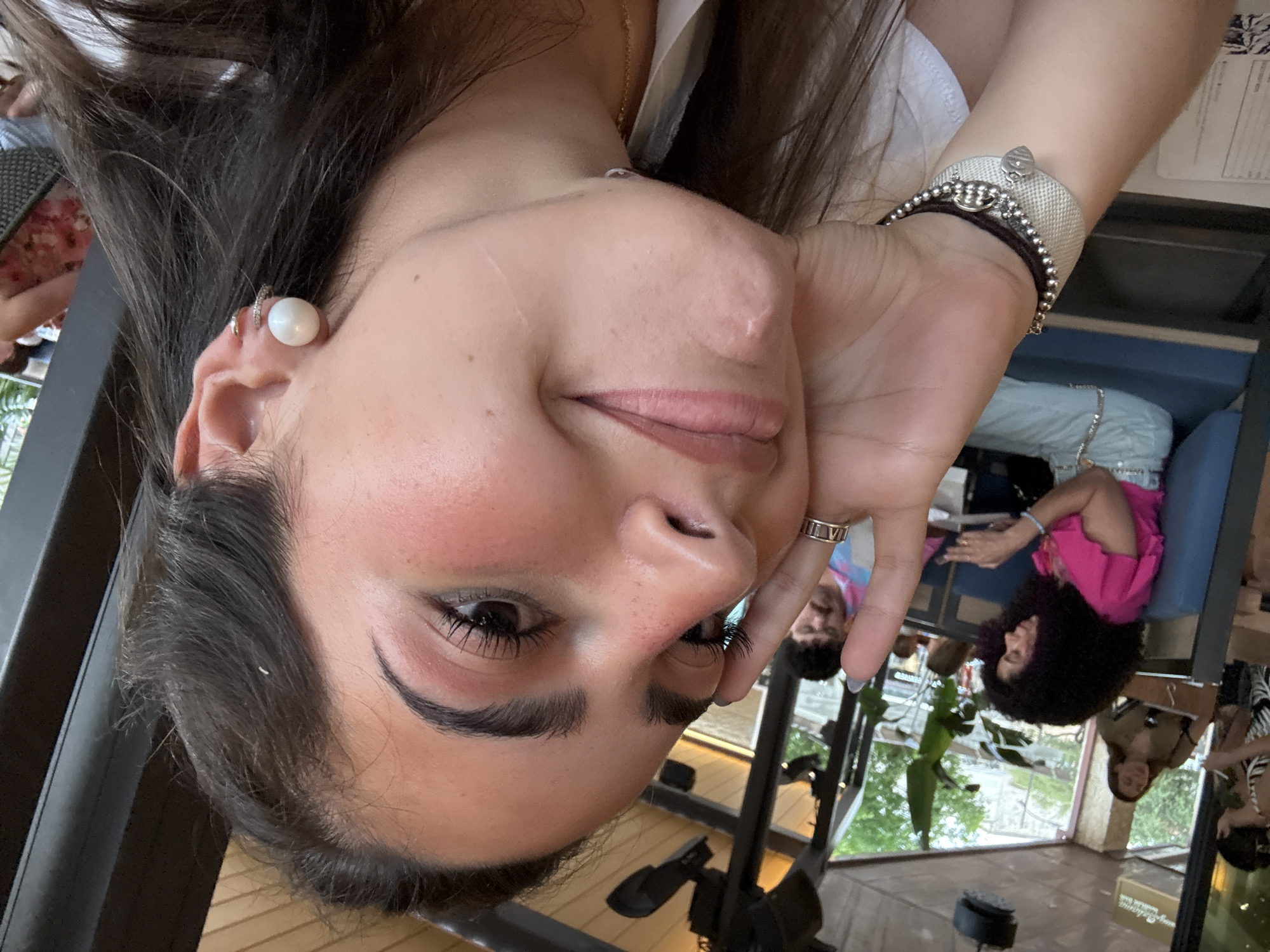A woman with dark hair is pictured in a close-up selfie. She wears a white pearl earring, a silver bracelet with charms, and a ring with roman numerals. She is smiling softly, with her hand near her face. In the background, there are other people seated at tables, including one wearing a bright pink shirt. Through the windows, greenery is visible. The scene appears to be inside a restaurant, possibly in San Antonio, United States of America. The tables and chairs suggest an indoor dining area.