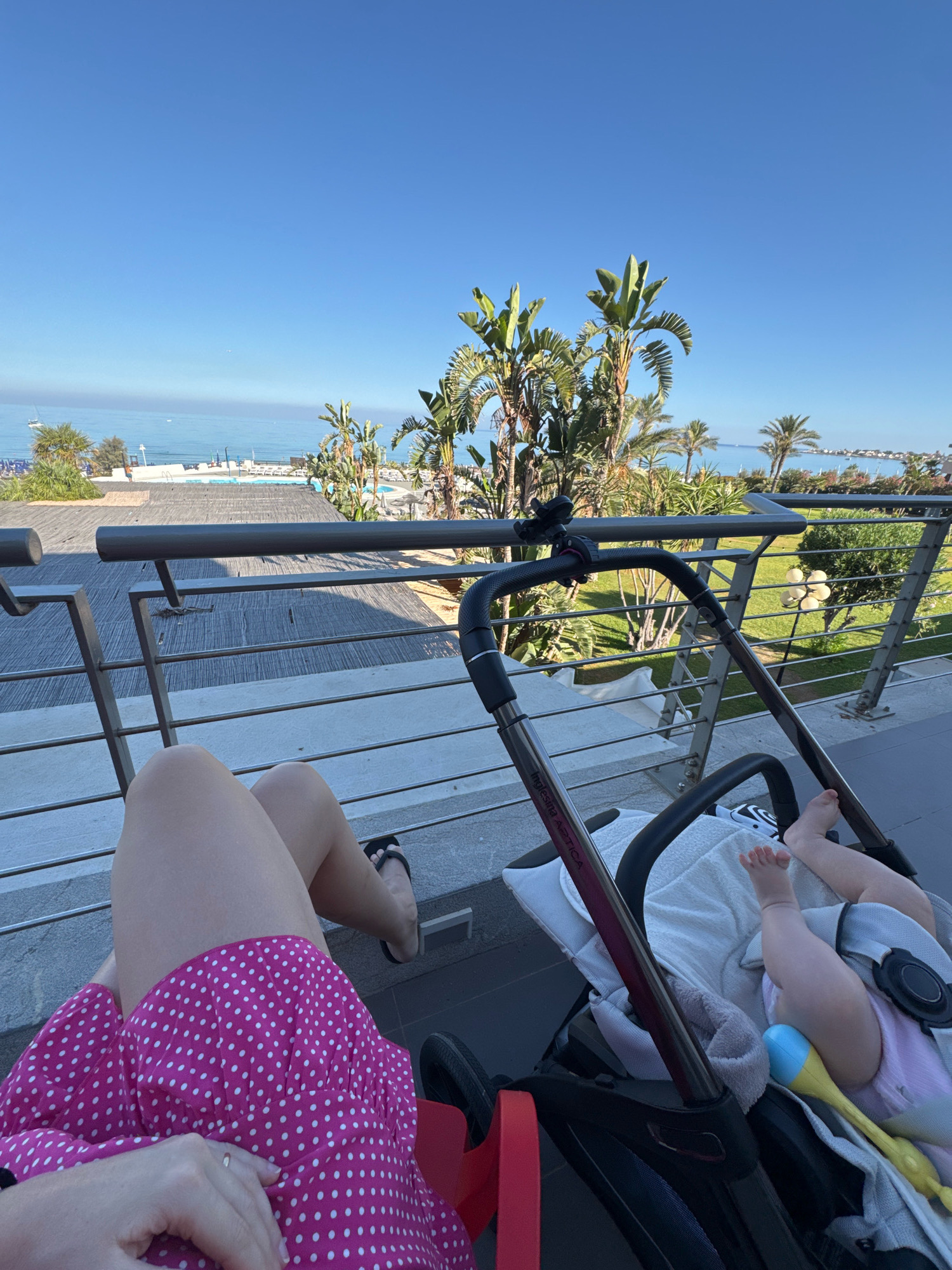 A person's legs, clad in a pink polka-dot dress and black flip-flops, are visible on the left side of the frame, resting on a balcony. In the foreground, a baby's feet and legs are seen in a stroller. The stroller, a dark color with red accents, sits on a balcony that overlooks a swimming pool and a body of water, likely the Mediterranean Sea, with palm trees and other foliage in the mid-ground. The sky is clear and blue. In the far distance, the coastline of Isola delle Femmine, Italy, with buildings and boats, is visible. The scene suggests a relaxed vacation or leisure setting.