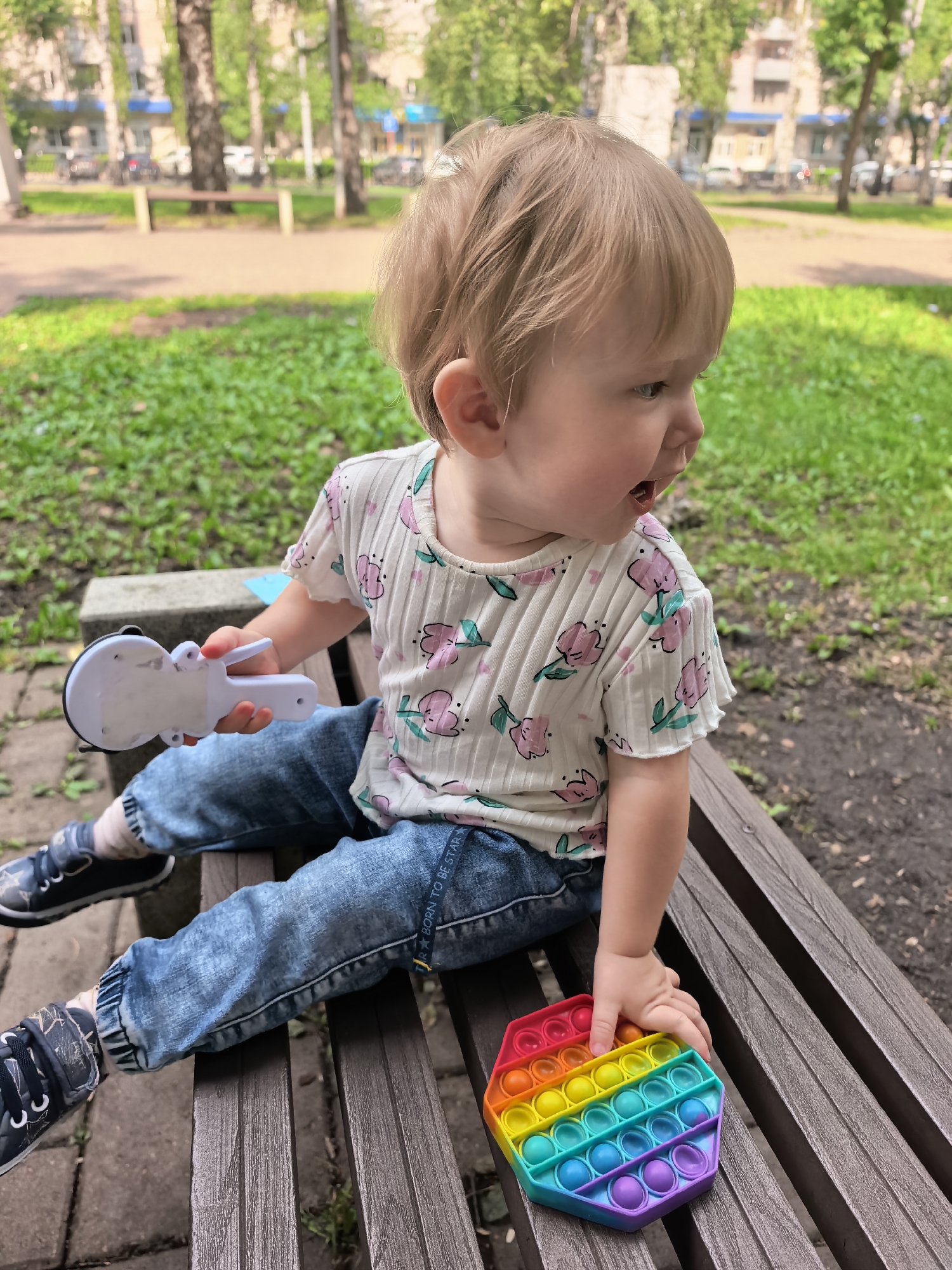 A young child, estimated to be between 1-2 years old, sits on a wooden park bench in Sterlitamak, Russia. The child has light brown hair and is wearing a white t-shirt with a floral pattern of pink flowers and green leaves, blue denim jeans, and dark blue sneakers with white laces and star patterns. The child is holding a white toy with a suction cup in their left hand and is touching a hexagonal rainbow-colored fidget toy with their right hand. The background shows green grass, trees, and buildings, suggesting an urban park setting. The lighting indicates it is daytime, with soft shadows. The overall scene depicts a moment of play in an outdoor recreational area.