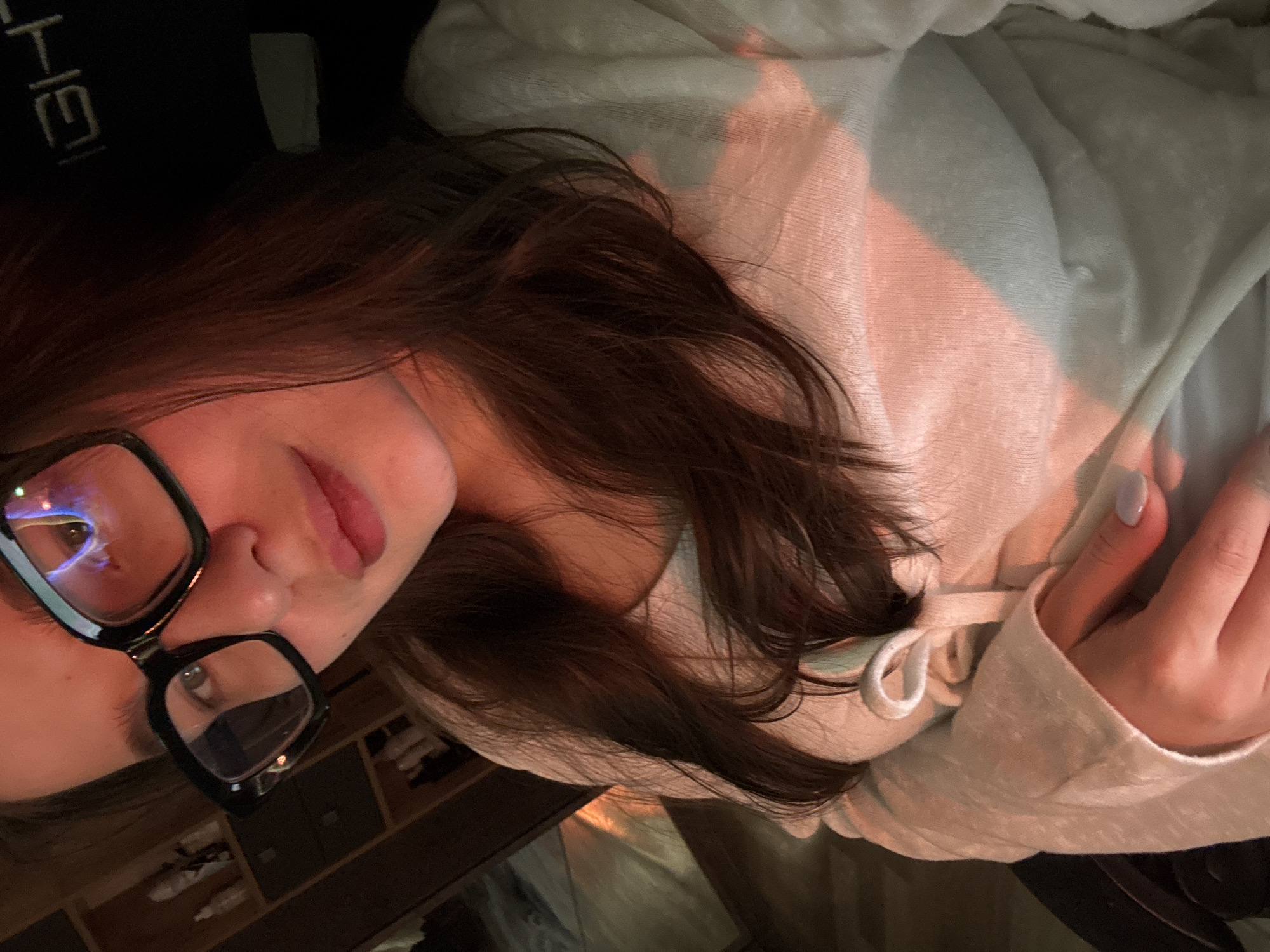 A woman with brown hair and black-rimmed glasses is shown from a close, tilted angle. She is wearing a light-colored sweatshirt. Light appears to be shining across her face and onto her clothing. In the background, there is a wooden shelf with objects on it and what appears to be a dark-colored headrest of a chair. There is no information available to identify the city as Chaikovsky, Russia.