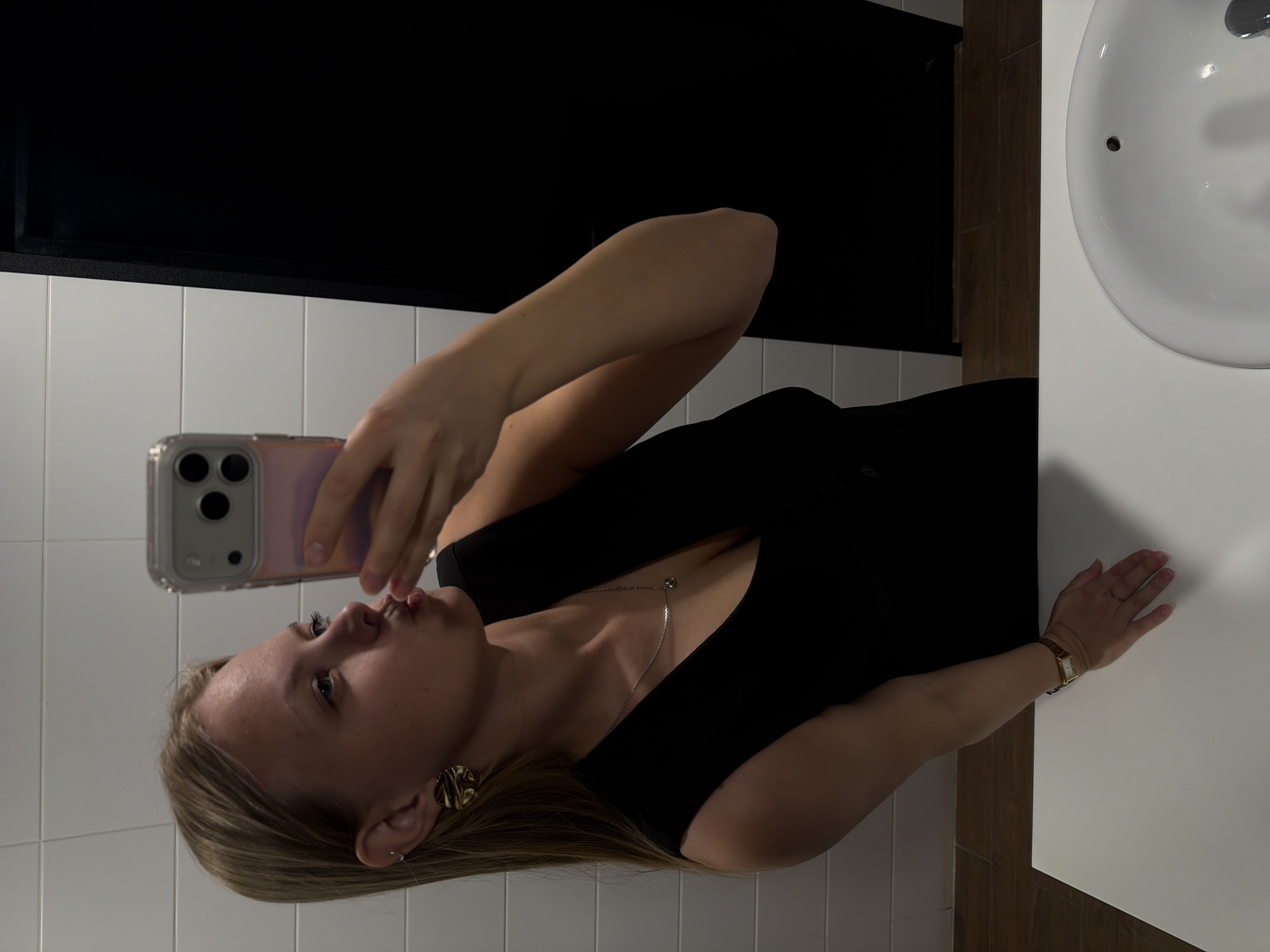 A woman is taking a selfie in a bathroom. The bathroom has white tiled walls and a black countertop. There is a white sink in the right of the image. The woman is wearing a black top and jewelry. She is holding a phone with a clear case. There is no information to suggest the city of Izhevsk, Russia as the location.