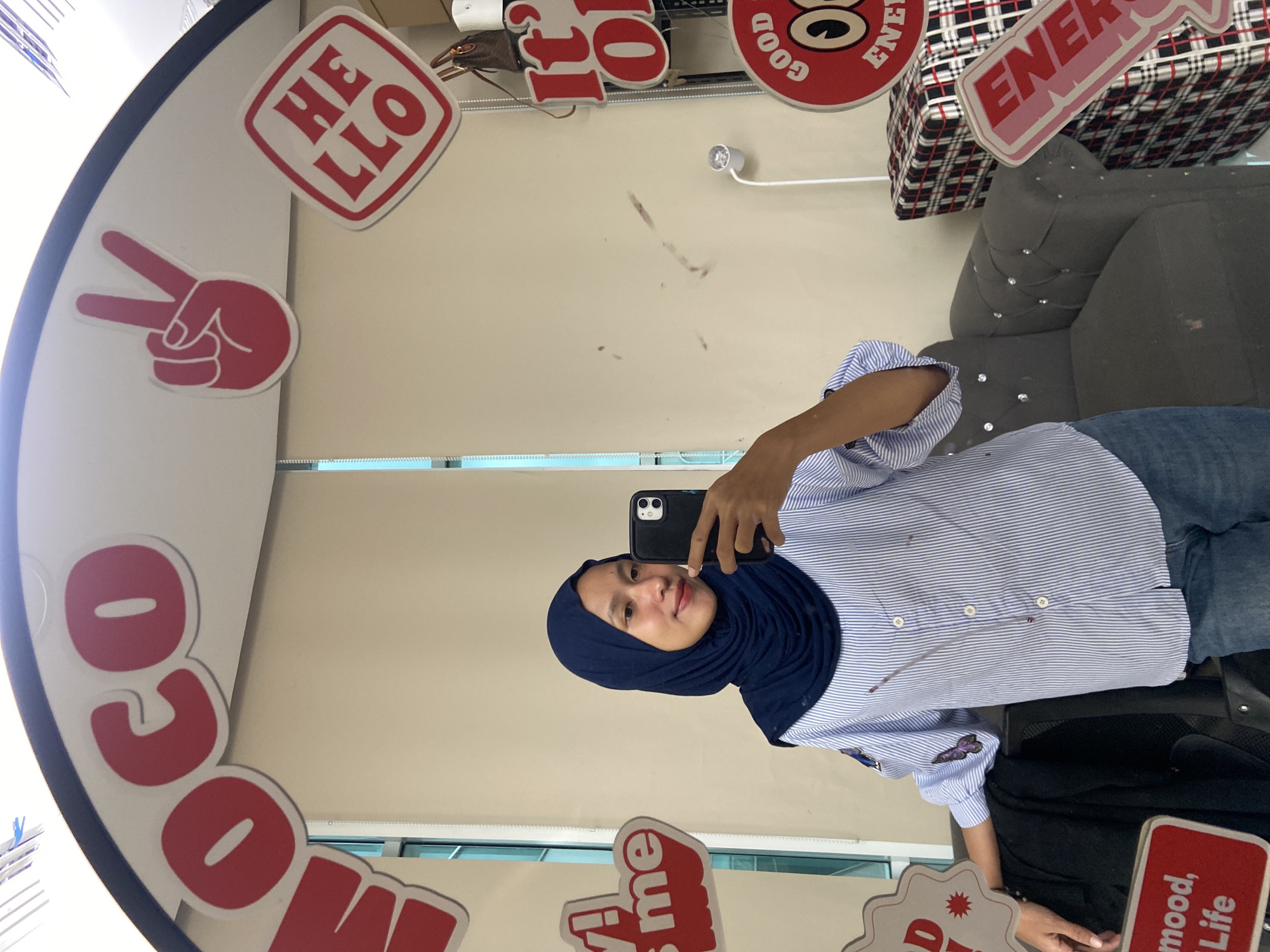 A woman wearing a blue hijab and a light blue striped shirt with blue jeans takes a selfie in a mirror. The mirror has motivational words on it. She is holding a black cellphone in her right hand. The location is North Jakarta, Indonesia.