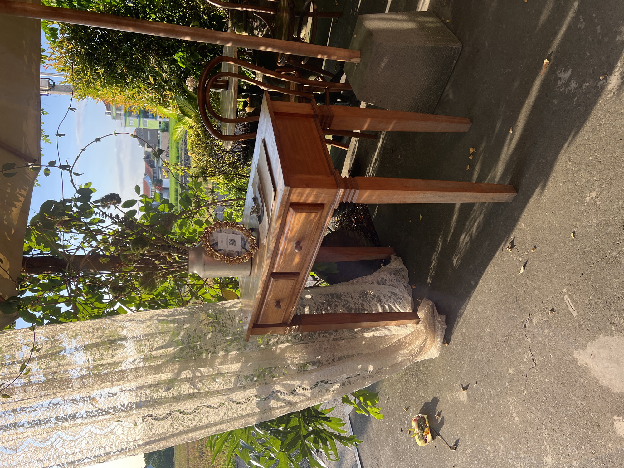The image depicts an outdoor scene, likely a patio or garden setting, located in North Kuta, Indonesia, during daylight hours. The primary objects in focus are a small wooden desk with two drawers, a vase containing leafy branches and a decorative frame, and a sheer, lace curtain partially obscuring the view. The desk is positioned on a concrete surface, casting shadows from surrounding elements. The scene is partially shaded by trees and an overhead structure, suggesting a sheltered outdoor area. In the background, lush greenery and hints of buildings can be observed, typical of the Indonesian landscape. The image suggests a staged or decorated area, possibly for an event or photoshoot.