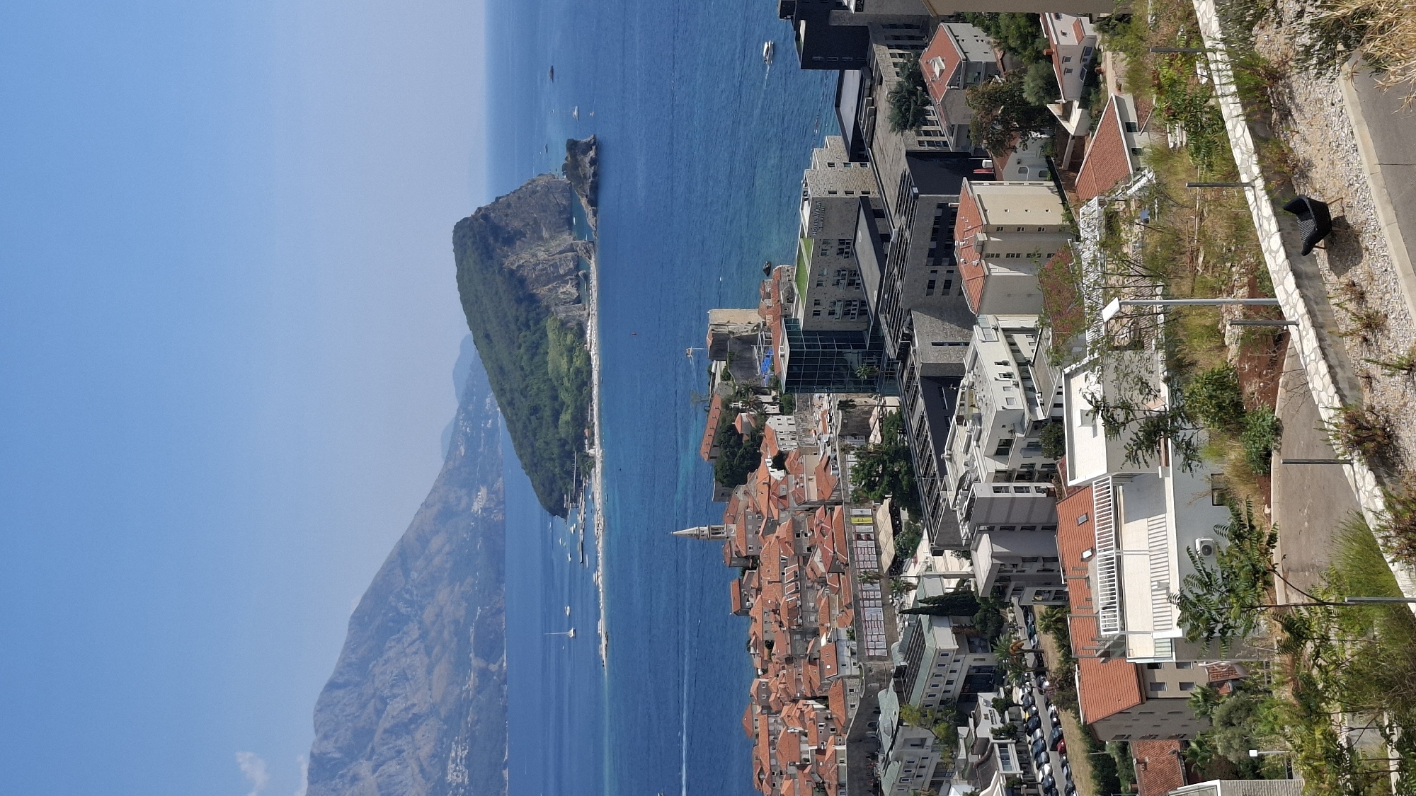 This is a high-angle, vertical, outdoor photograph of the city of Budva, Montenegro, on a clear, sunny day. The foreground shows a terraced area with dry vegetation, a paved path, a stone wall, and a dark armchair. In the mid-ground, a cluster of buildings with orange-tiled roofs and white walls dominates, with a prominent bell tower from a church. Further back, modern buildings, some with glass facades and green roofs, are visible. The background features a large, verdant island connected by a sandbar to a beach area, with the blue Adriatic Sea surrounding it. In the distance, hazy mountains rise against a bright blue sky. Several boats are scattered on the water, including sailboats and smaller vessels. The scene depicts a coastal city with a mix of old and new architecture, set against a beautiful natural backdrop.