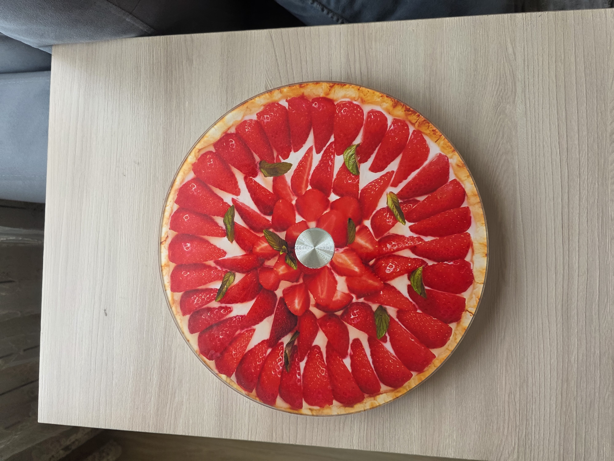 A circular strawberry tart, depicted in a high-quality photograph, is centrally placed on a light-colored wooden table. The tart is adorned with numerous sliced strawberries, arranged in a radial pattern, with some mint leaves interspersed among them. The crust of the tart is visible at the edge, appearing golden brown. In the center of the tart, a silver metallic element with a spiral pattern is present. The table surface exhibits a subtle wood grain texture. To the right of the table, a portion of a grey upholstered chair is visible, and further to the right, dark fabric, possibly denim, can be seen. The background includes a blurred view of vertical blinds. The overall scene is well-lit, suggesting an indoor setting. The image does not contain any identifiable landmarks or elements that would place it specifically in Скалба Снт, Russia.