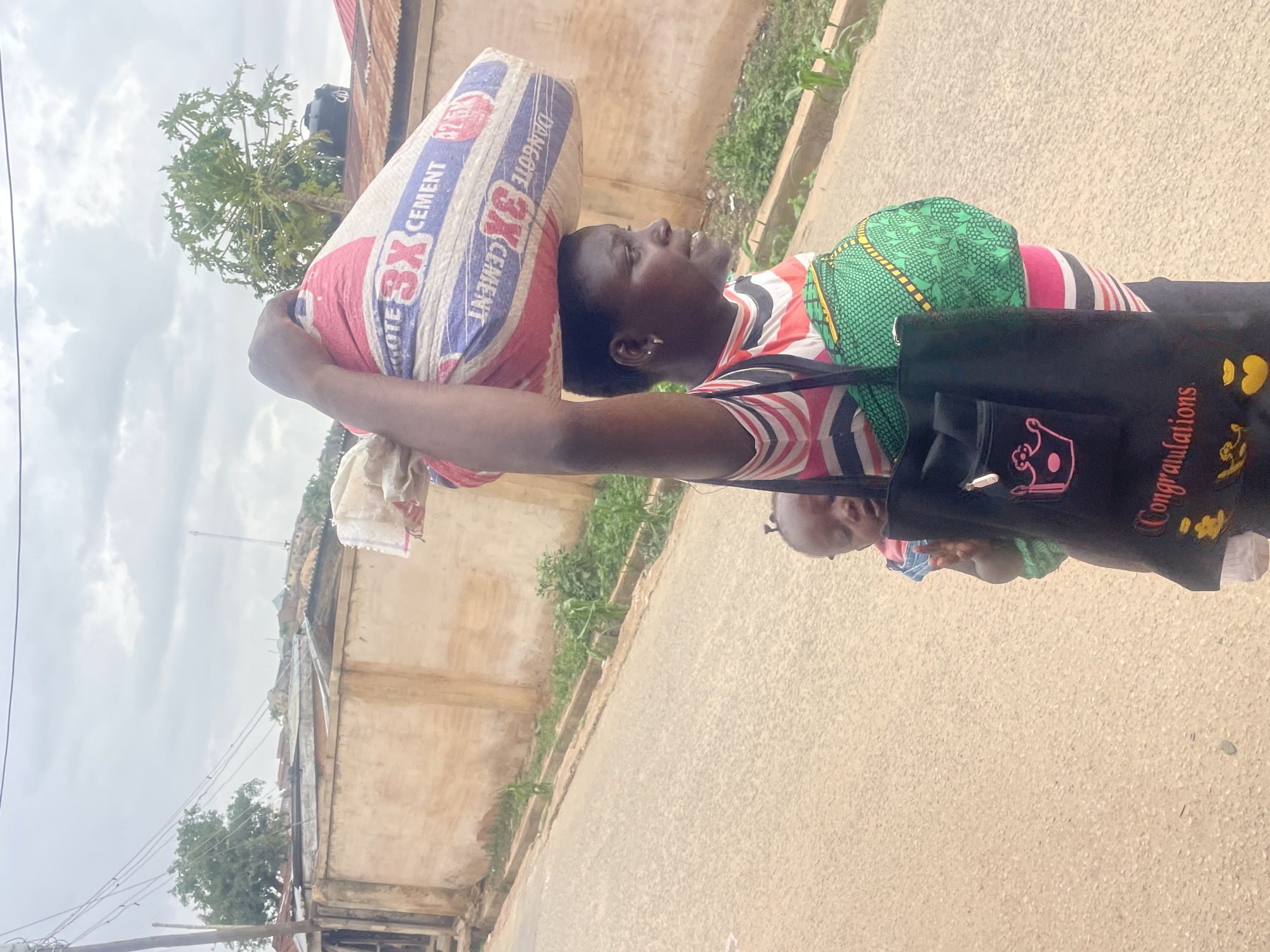 A woman is carrying a large bag of Dangote 3X cement on her head while walking on a paved road in Jos, Nigeria. She has a baby wrapped on her back with a green patterned cloth and is carrying a black bag with "Congratulations" printed on it over her shoulder. The buildings lining the road are simple, with visible wear and tear, and there is vegetation growing nearby. The sky is overcast.