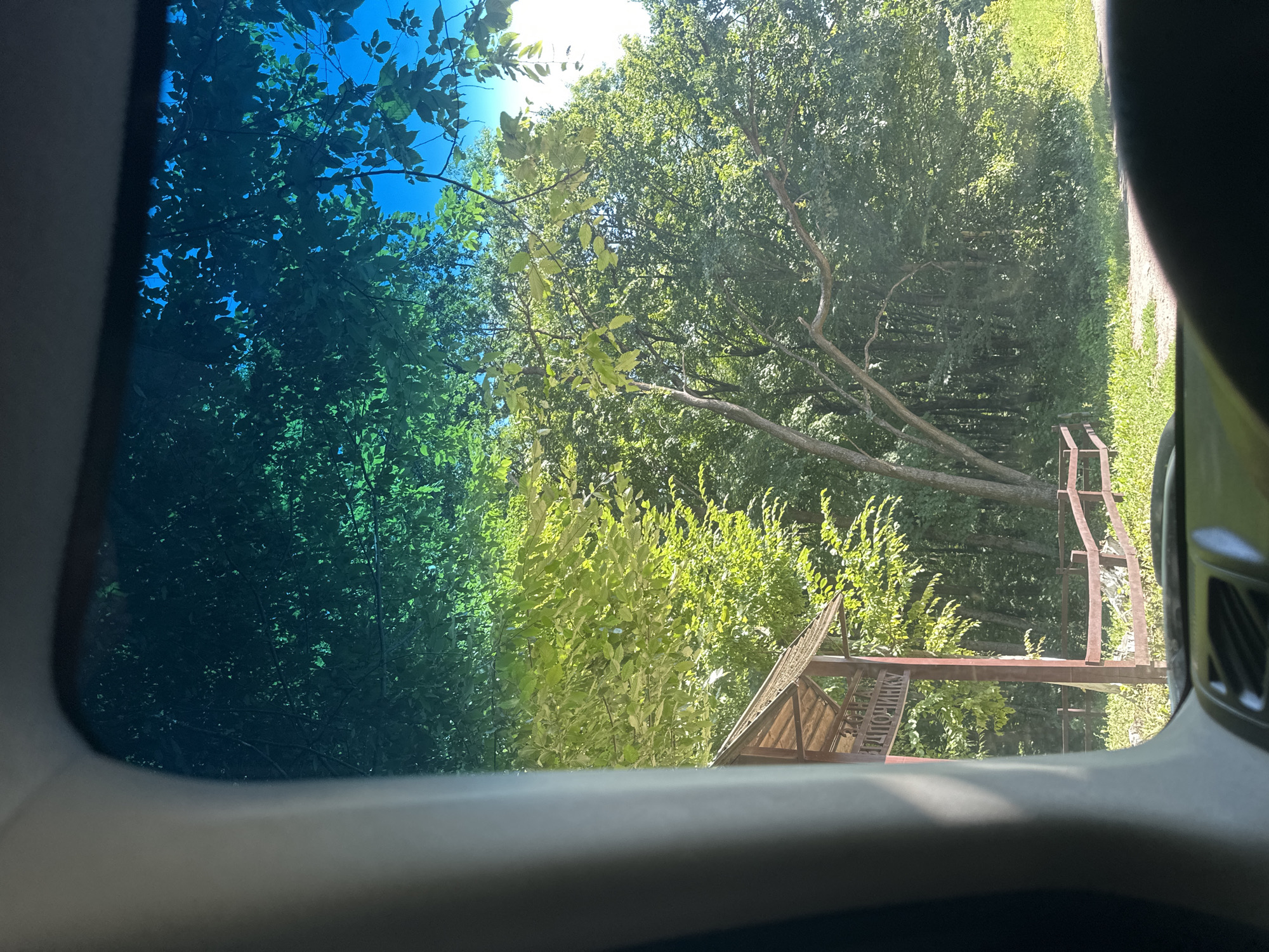 The image captures a view from inside a vehicle looking out through the windshield. The windshield has a blue tint, particularly noticeable towards the top, which casts a greenish hue on the foliage. In the foreground, the interior of the car is visible, including a portion of the dashboard and a dark, curved object, possibly the steering wheel or dashboard.
Outside the car, a wooden structure with a slanted roof is partially visible on the left side of the frame. This structure appears to be a signpost or an entrance gateway, with some text written on it that is partially obscured. A brown wooden fence runs horizontally across the middle ground of the image. Behind the fence, lush green trees dominate the scene, with sunlight filtering through the leaves, creating bright spots and dappled shadows. The sky above is a clear blue, with some branches and leaves silhouetted against it. The overall environment suggests an outdoor setting, likely a natural area or a park, given the presence of trees, grass, and the rustic fence and signage. The context provided places this scene in Shatava, Ukraine.