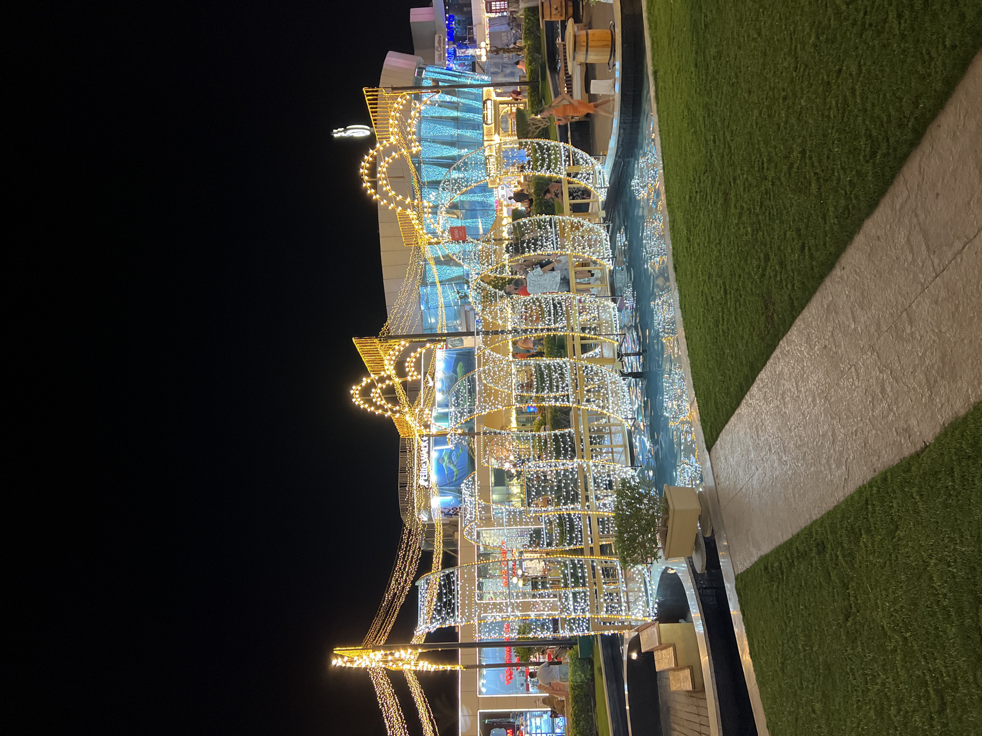 A nighttime exterior shot captures a brightly lit pathway leading into a fountain area. A woman in an orange dress is seen standing near a wooden barrel table in the foreground. Behind her, a decorative structure illuminated with white lights resembles a boat. Beyond the fountain, a building with large blue windows is adorned with cascading strings of yellow lights. Several people are visible in the background, walking and interacting near shop entrances. The scene appears to be set in a pedestrianized area within Sharm El Sheikh, Egypt, likely a commercial or entertainment district given the architectural style and illuminated displays.