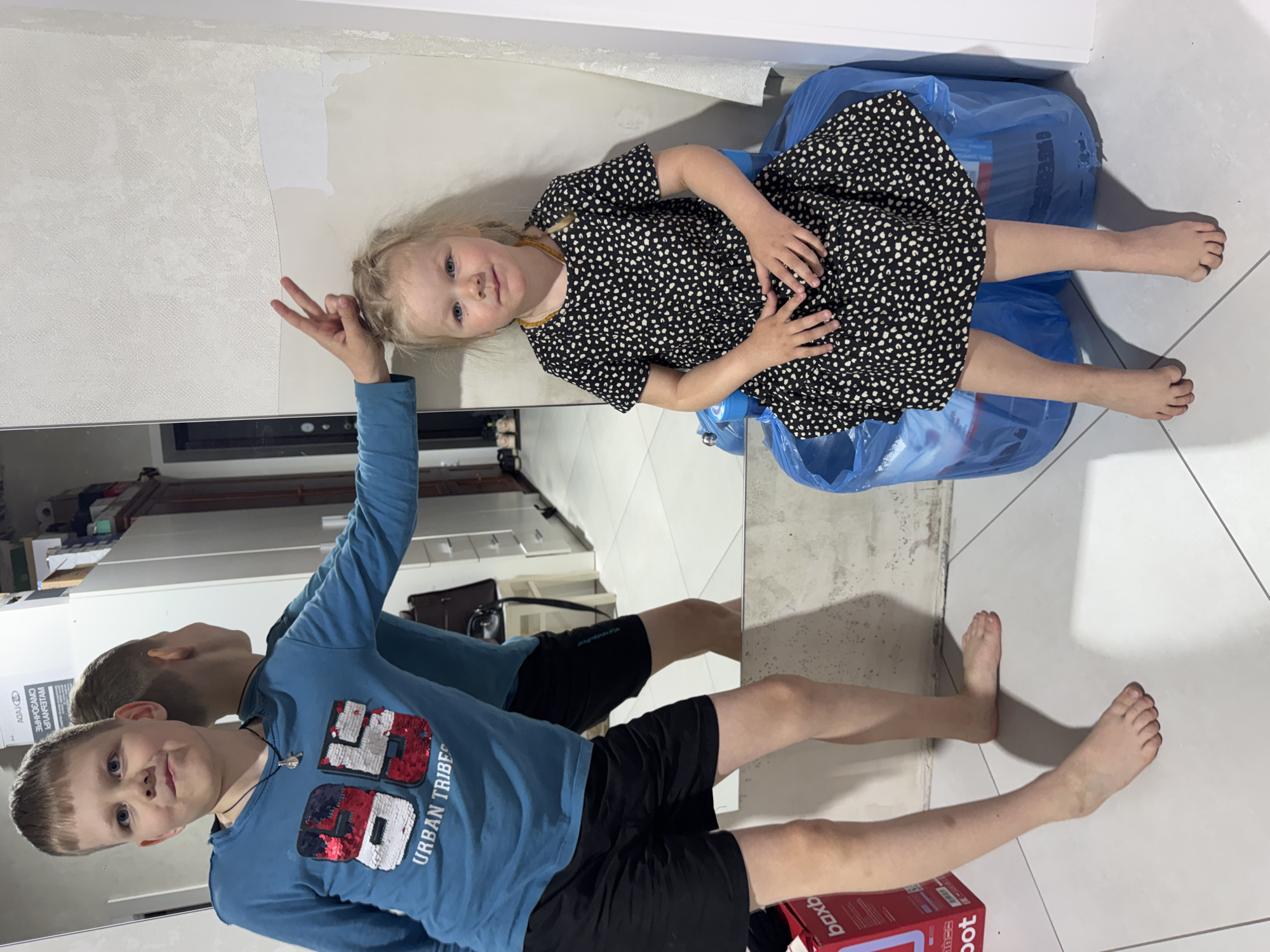 A boy stands in front of a mirror, striking a pose with two fingers raised in a "peace" sign. He wears a long-sleeved blue shirt with a sequined "86" and "URBAN TRIBES" on it, paired with black shorts. Reflected in the mirror is his image. Next to him is a young girl wearing a black dress with white polka dots, sitting on a blue plastic garbage bag. The background suggests an indoor setting with white walls, possibly a home interior with elements of renovation, as there are remnants of wallpaper peeling off. A red box is visible in the lower right corner. Location is potentially in an area near Gornyi (Dvurechenskoe S/P), Russia.
