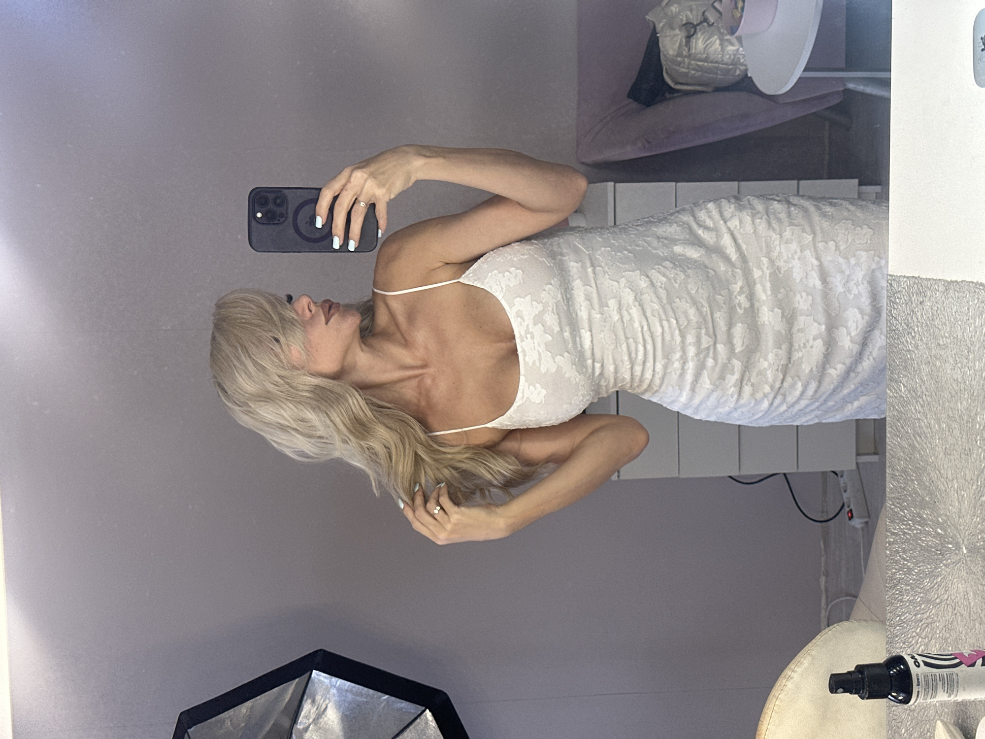 A woman is taking a mirror selfie in a dimly lit room. She has long, blonde hair and is wearing a white, textured dress. She is holding a black smartphone in her right hand to capture the photo. Her left arm is extended, and she is touching her hair. There's a ring on her left ring finger.
The background of the room appears to be a living space. A blurred image of a sofa with a pillow is visible in the upper right corner. A lamp with a light-colored shade is also visible. In the lower part of the frame, part of a light fixture or umbrella is seen. The walls are a muted gray color. The overall ambiance suggests an indoor setting, possibly a home or a studio. The lighting is soft, and the image has a vertical orientation as if the camera was held in portrait mode, but the entire scene is rotated. The image is a reflection in a mirror, so it shows the room from a reversed perspective.
There is no discernible time of day or weather information. The emotion conveyed is neutral, focused on the act of taking a selfie. No text is clearly visible in the image.