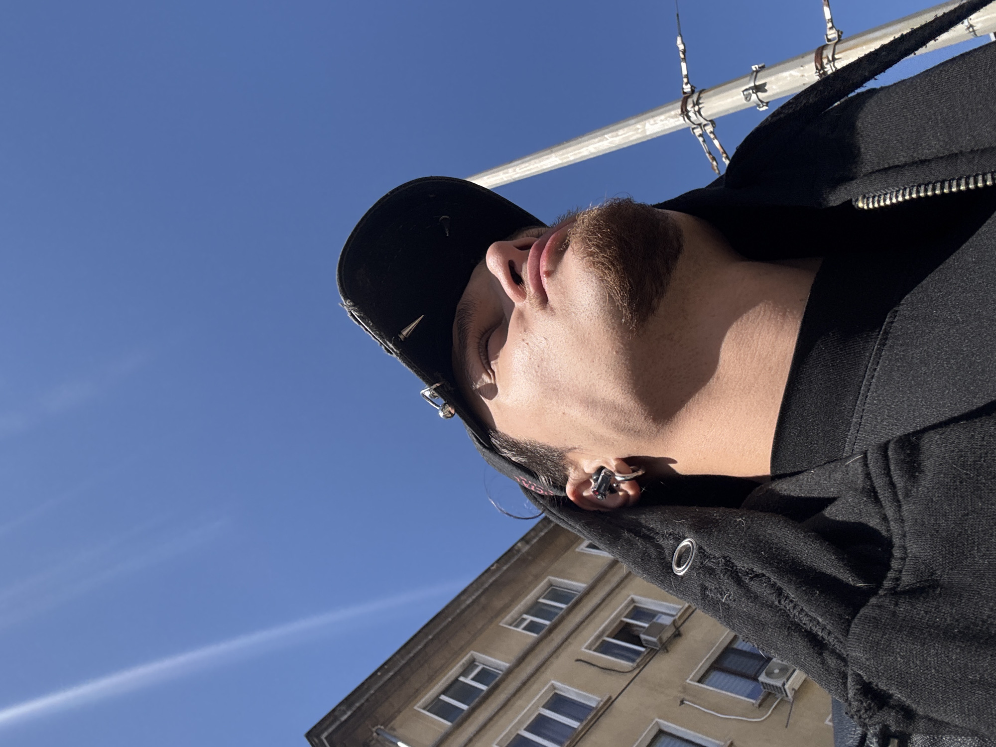 A low-angle, rotated shot captures a fair-skinned individual wearing a black baseball cap and jacket against a bright blue sky. The cap has a small, possibly metal, detail on its side. The person's head is tilted back, exposing their neck and a light-brown beard. They have several piercings in their ear. A metal pole or pipe with clamps and a chain is visible above them. In the lower portion of the frame, part of a beige, multi-story building with white-framed windows and air conditioning units attached to some windows can be seen. The building's architecture suggests an urban environment. Based on the architectural style and language, the building is likely located in the city of Sofia, Bulgaria.