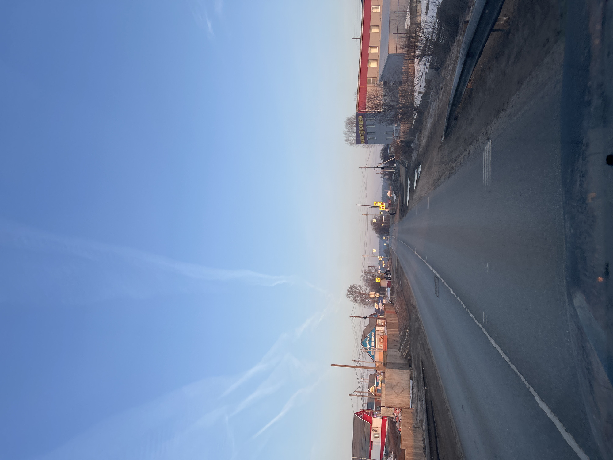 The image depicts a road in the city of Гаревая, Russia. The road stretches into the distance, flanked by buildings and fences on both sides. The sky is clear and blue with contrails. A building with a red roof is on the right side of the road. There is also a building with a sign in Russian. A metal guardrail is on the right side. There are some utility poles along the road.