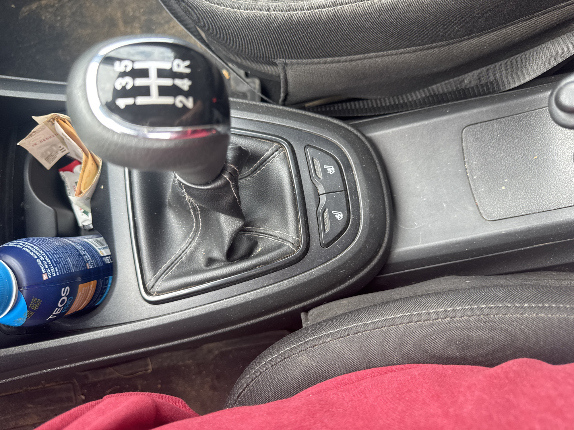 The image shows the interior of a car, focusing on the gear shifter and the center console. A red piece of fabric, likely clothing, is visible on the left side of the image, draped over a seat. The gear shifter has markings for gears 1, 3, 5, 2, 4, and R. Below the gear shifter, there are two buttons with icons resembling heated seats. To the right of the gear shifter, a cup holder contains a blue bottle with the brand name "TEOS PRO" visible, and a crumpled piece of paper. The overall environment appears to be a car's interior, possibly parked or in motion. There are no visible people in the image. The location is specified as the city of Chastyye, Russia.