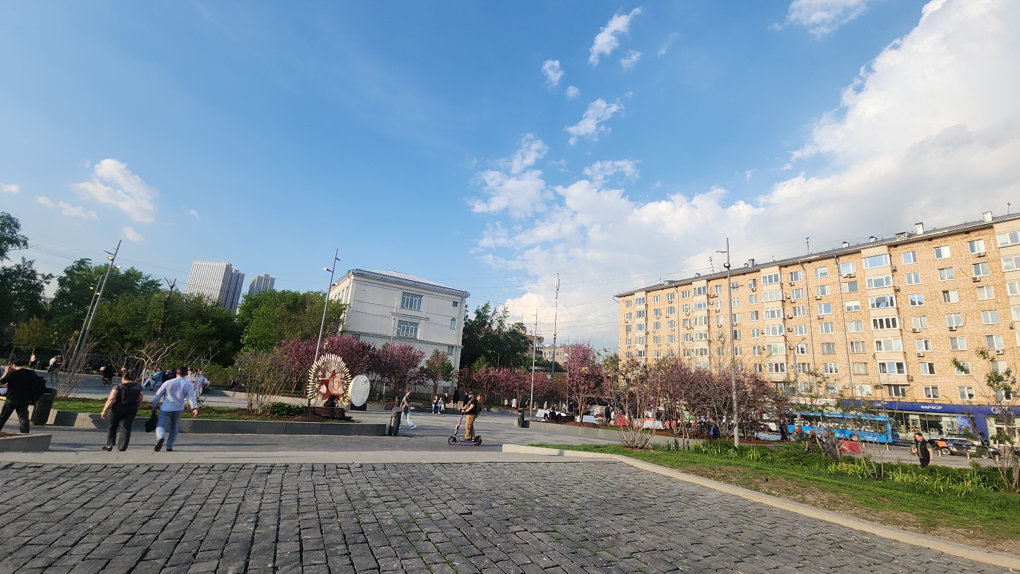 The image depicts an urban scene, likely a public square in Moscow, Russia. The foreground features a paved area with cobblestones leading to a concrete platform. Several people are present: a couple walking, an individual on a scooter, and others scattered throughout the scene. There are benches, planters with trees displaying pink blossoms, and decorative art installations. In the background, buildings of varying sizes and architectural styles are visible, along with some modern high-rise buildings. A blue bus is seen driving along the road behind the green patch. The sky is blue with scattered clouds, indicating a clear day.