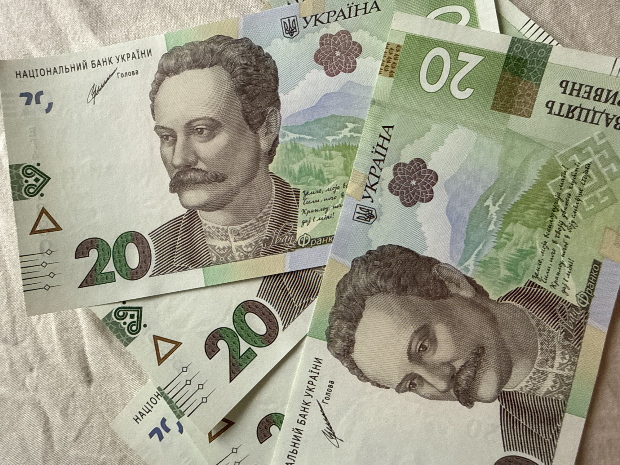 The image displays a close-up, overhead view of several Ukrainian 20 hryvnia banknotes. The banknotes are fanned out and overlapping on a textured, light-colored surface, possibly fabric. The details of the banknotes are clearly visible, including portraits of Ivan Franko, national symbols, and the denomination "20". The lighting is somewhat uneven, casting subtle shadows. The background does not provide any information about the city of Bad Sachsa, Germany.