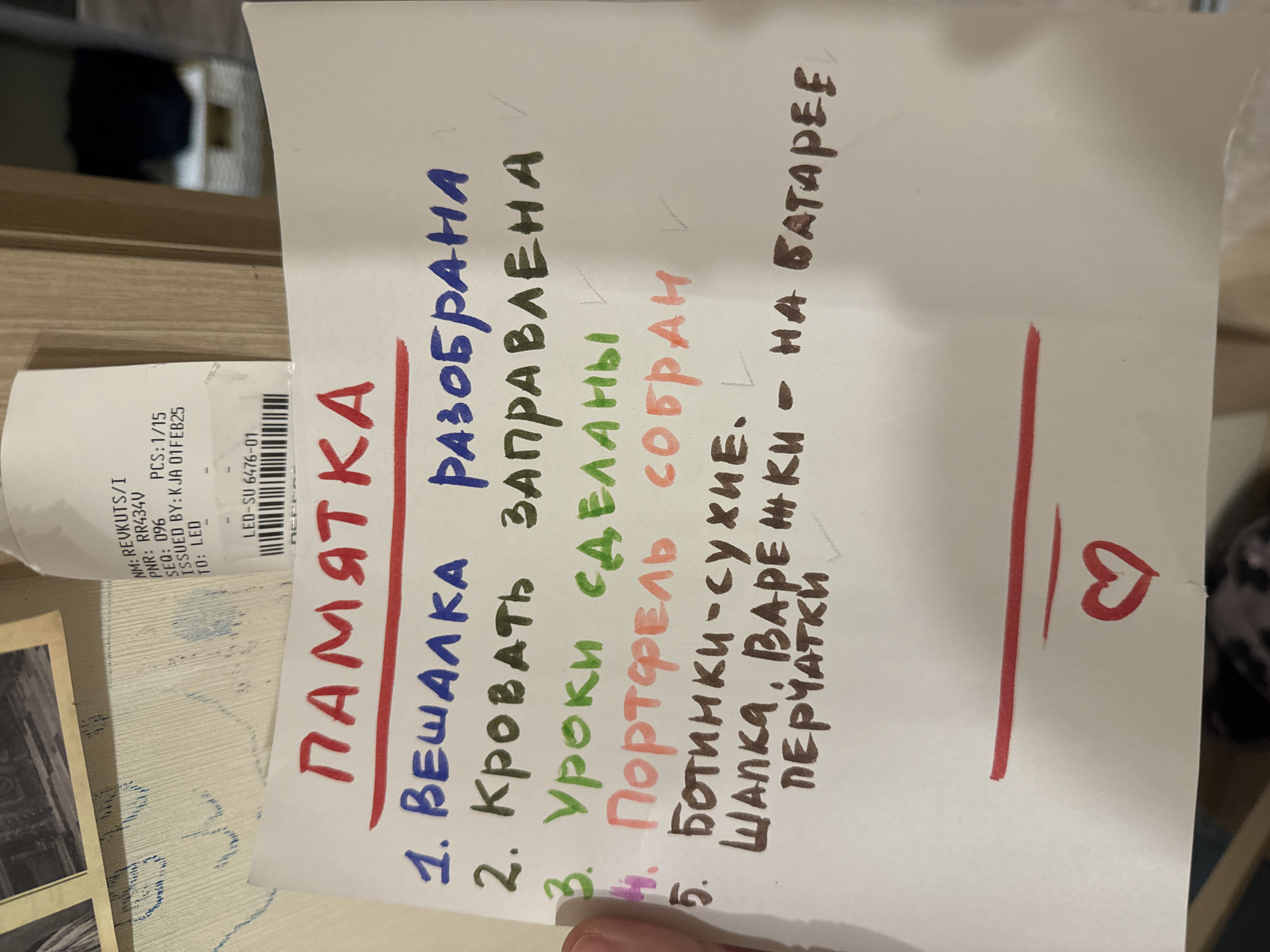 The image displays a handwritten reminder note, titled "ПАМЯТКА" (MEMO / REMINDER) in red and underlined, positioned on a light-colored wooden surface. A human thumb is visible at the bottom right, suggesting the note is being held or has just been placed. To the left of the note, a partially visible paper receipt with printed text and a barcode is discernible. The receipt includes details such as "NM: REVKUTS/I", "PNR: RR434V", "SEQ: 096", "PCS:1/15", "ISSUED BY: K JA 01FEB25", and "LED-SU 6476-01", indicating a recent purchase or transaction.
The note itself is a checklist of five tasks, written in Russian with various colored pens, and features checkmarks next to several items, signifying completion. The tasks are:
1. "ВЕШАЛКА РАЗОБРАНА" (HANGER DISASSEMBLED / COAT RACK TAKEN APART), written entirely in blue.
2. "КРОВАТЬ ЗАПРАВЛЕНА" (BED MADE), with "КРОВАТЬ" in black and "ЗАПРАВЛЕНА" in green.
3. "УРОКИ СДЕЛАНЫ" (HOMEWORK DONE), both words written in green.
4. "Портфель Собран" (BACKPACK PACKED), both words written in pink.
5. "БОТИНКИ-СУХИЕ. ШАПКА, ВАРЕЖКИ - НА БАТАРЕЕ" (BOOTS - DRY. HAT, MITTENS - ON THE RADIATOR), all written in brown.
These tasks suggest a daily routine, possibly for a child or someone getting ready for the day, with an emphasis on personal organization and preparing for cold weather (drying boots, hat, and mittens on a radiator). The variety of pen colors adds a personal and caring touch. At the bottom right corner of the note, a small red heart is drawn, implying affection or a gentle reminder from a loved one, likely a parent. The setting is an indoor environment, likely a home in Krasnoyarsk, Russia, given the Cyrillic script and the provided context.
