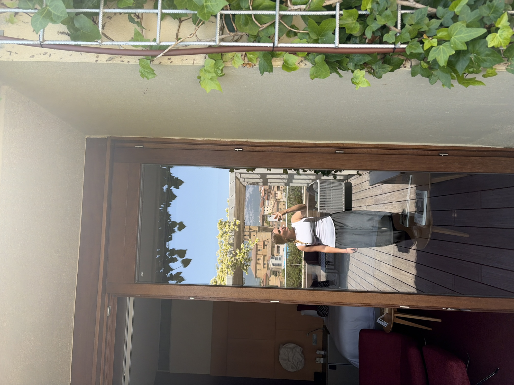 A reflection of a person with a mobile phone takes a picture of a city landscape from a balcony through a window. The person is wearing a white top and a gray skirt. The city landscape features buildings, trees, and mountains in the distance under a clear blue sky. Inside the room, a red sofa and a white bed can be seen. The window frame is made of wood. Green vines are visible above the window frame.