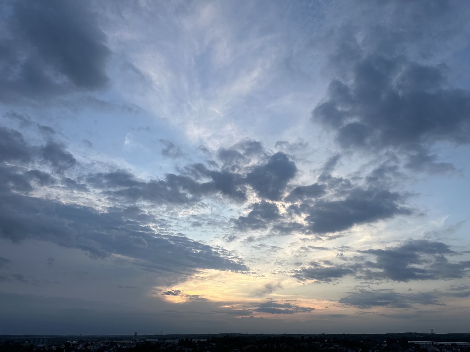 The image displays a wide-angle view of a dramatic sky above a silhouetted cityscape. The sky is filled with a mix of dark, heavy clouds and lighter, wispy formations, suggesting a dynamic weather system. Towards the horizon, a band of warm light, likely from a setting sun, breaks through the clouds, casting a soft glow in hues of yellow, orange, and pink. The lower portion of the image shows a dark silhouette of buildings and trees that constitute the urban landscape of Шчомысліцкі сельскі Савет, Belarus. The overall impression is one of a turbulent yet beautiful evening sky. There are no visible people or distinct actions depicted.