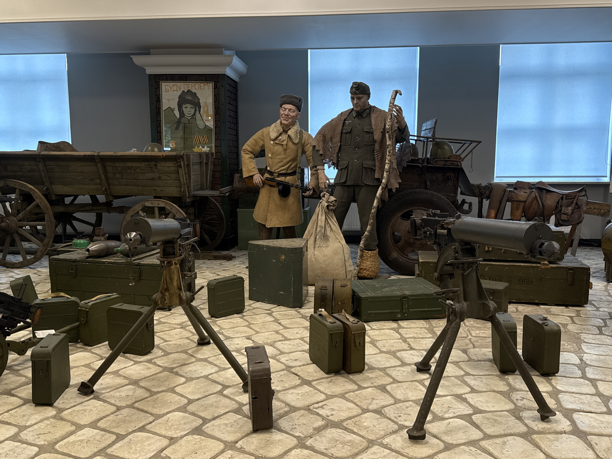 The image depicts a museum exhibit in Arkhangel’skoye, Russia, showcasing military artifacts. Two mannequin figures, dressed in historical military attire, stand amidst a collection of weaponry and equipment. The figure on the left wears a tan coat and cap, holding a rifle. The figure on the right is in a green uniform with a cap, holding a wooden stick. Surrounding them are various items including a wooden wagon, a cannon on a tripod, ammunition boxes, a sack, and a poster depicting a young woman in military uniform with the text "БУДУ ГЕРОЕМ!" (I WILL BE A HERO!). The floor is tiled with large, irregular stones. The background features blue-tinted window coverings and a section of a building with brick detailing. The exhibit appears to be indoors.