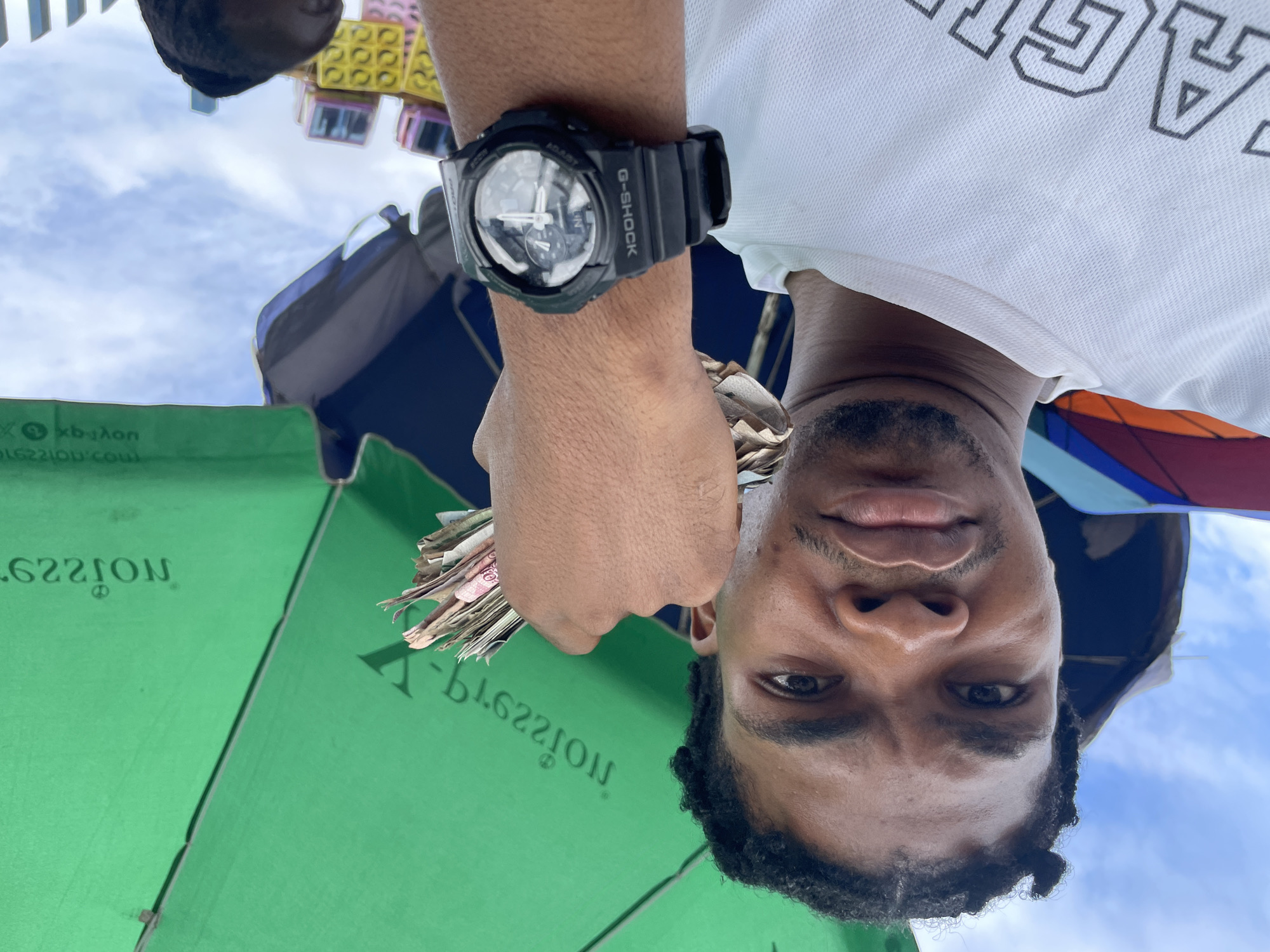A man, identified as being in Ajah, Nigeria, is shown from a low-angle perspective. He is holding a fan of banknotes in his right hand, which is adorned with a black G-SHOCK watch. The man is wearing a white t-shirt. He is partially obscured by a large green umbrella, which has text printed on it. In the background, there are some colorful structures that appear to be toys, and a cloudy sky. The overall impression is of an outdoor scene, possibly a market or a vendor's stall, given the presence of the umbrella and the man's attire.