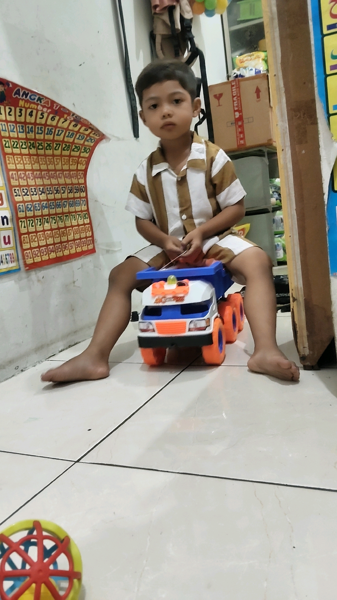 A young boy of Indonesian descent is sitting on a tiled floor, playing with a toy truck. The boy is wearing a short-sleeved shirt with vertical brown and white stripes and matching shorts. He is looking directly at the camera with a neutral expression. The toy truck is blue and white with orange accents and four orange wheels. In the background, there is a wall with a chart displaying numbers and letters, suggesting an educational setting or a child's room. There are also some boxes and shelves visible, along with what appear to be balloons hanging from the ceiling. The flooring is light-colored tile with dark grout lines. The scene appears to be indoors in a residential or educational environment within Mekarmukti, Indonesia.
