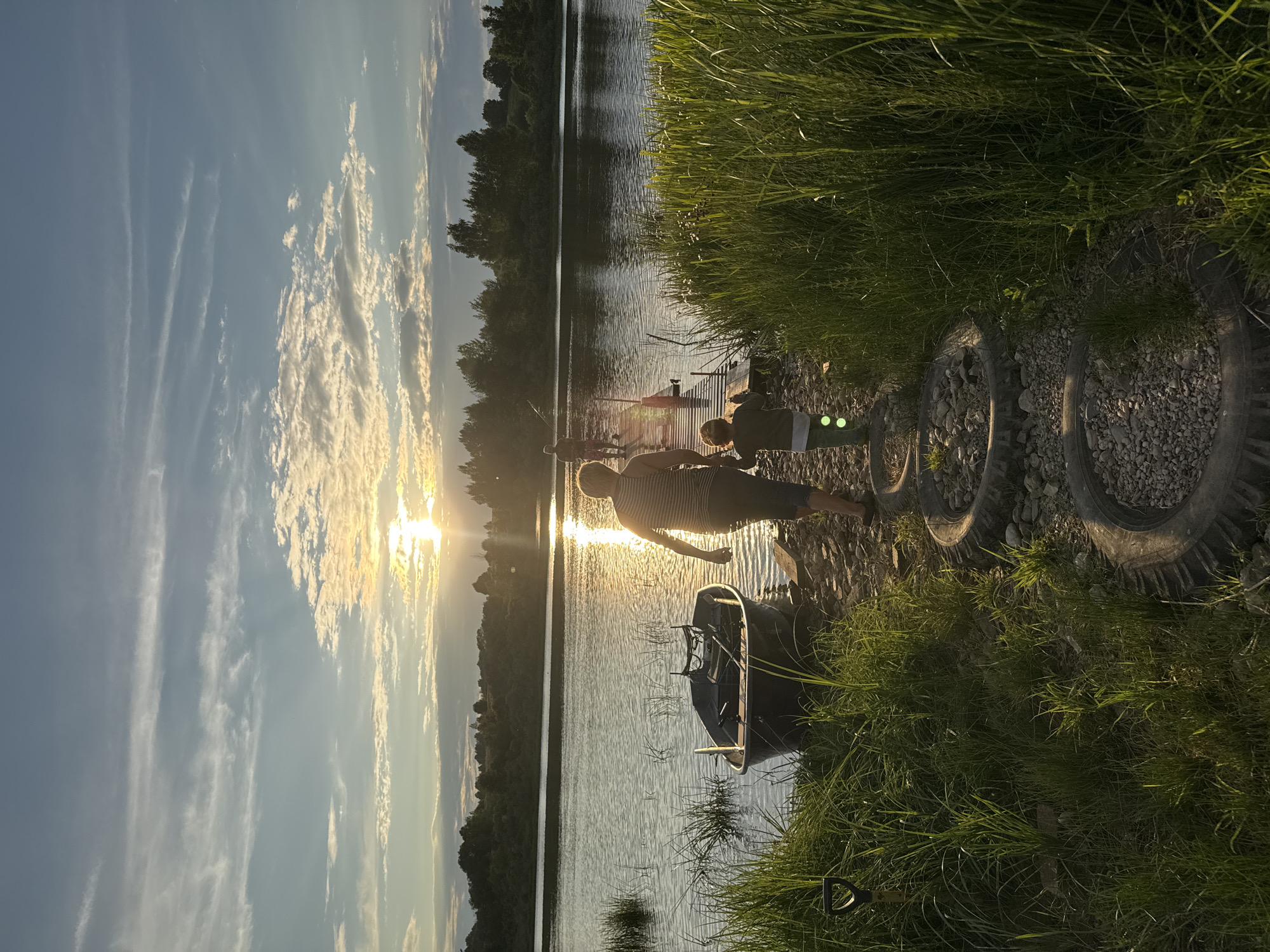 This is a full, outdoor, vertical, eye-level shot of a riverbank scene at dusk. The sun is setting to the left, casting a warm glow on the water and sky. The sky is a mix of blue and white clouds, with streaks of cirrus clouds high up.
In the foreground, tall green grass lines the right side of the frame. A pathway made of old tires filled with gravel leads from the bottom of the image towards a wooden pier. A shovel is stuck in the ground to the left of the pathway.
On the left side of the image, a small, dark-colored boat is moored by the bank. The water is calm with ripples reflecting the sunlight.
On the wooden pier, two people are visible. An adult, silhouetted against the setting sun, walks away from the camera towards the water. They are wearing a striped tank top and dark shorts. A child, also silhouetted, walks slightly ahead of the adult, holding their hand. The child is wearing a green shirt and pants. Further down the pier, another person is visible, possibly fishing, as they are holding a fishing rod.
The background consists of a dense line of trees and foliage along the opposite bank of the river. The overall atmosphere is peaceful and serene, capturing a moment of outdoor activity at sunset. The location is identified as Sitno, Russia.