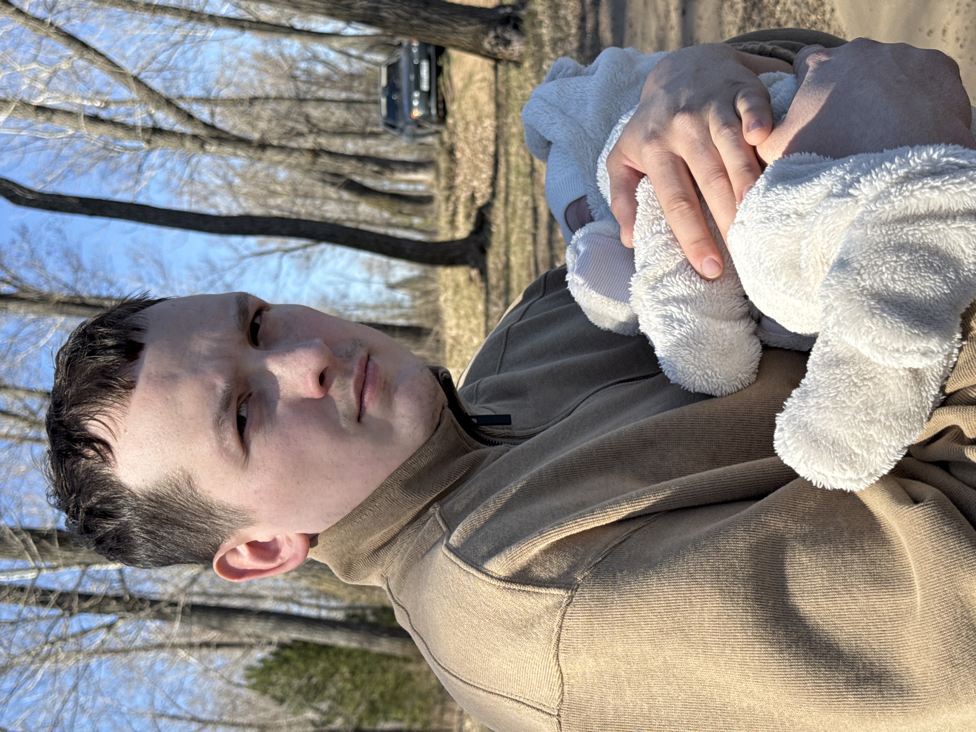 A man holds a baby wrapped in a white, fluffy blanket outdoors. The man is wearing a brown hooded sweatshirt. The background consists of trees with bare branches and a vehicle is visible in the distance. The photo was taken in Chaikovsky, Russia.
