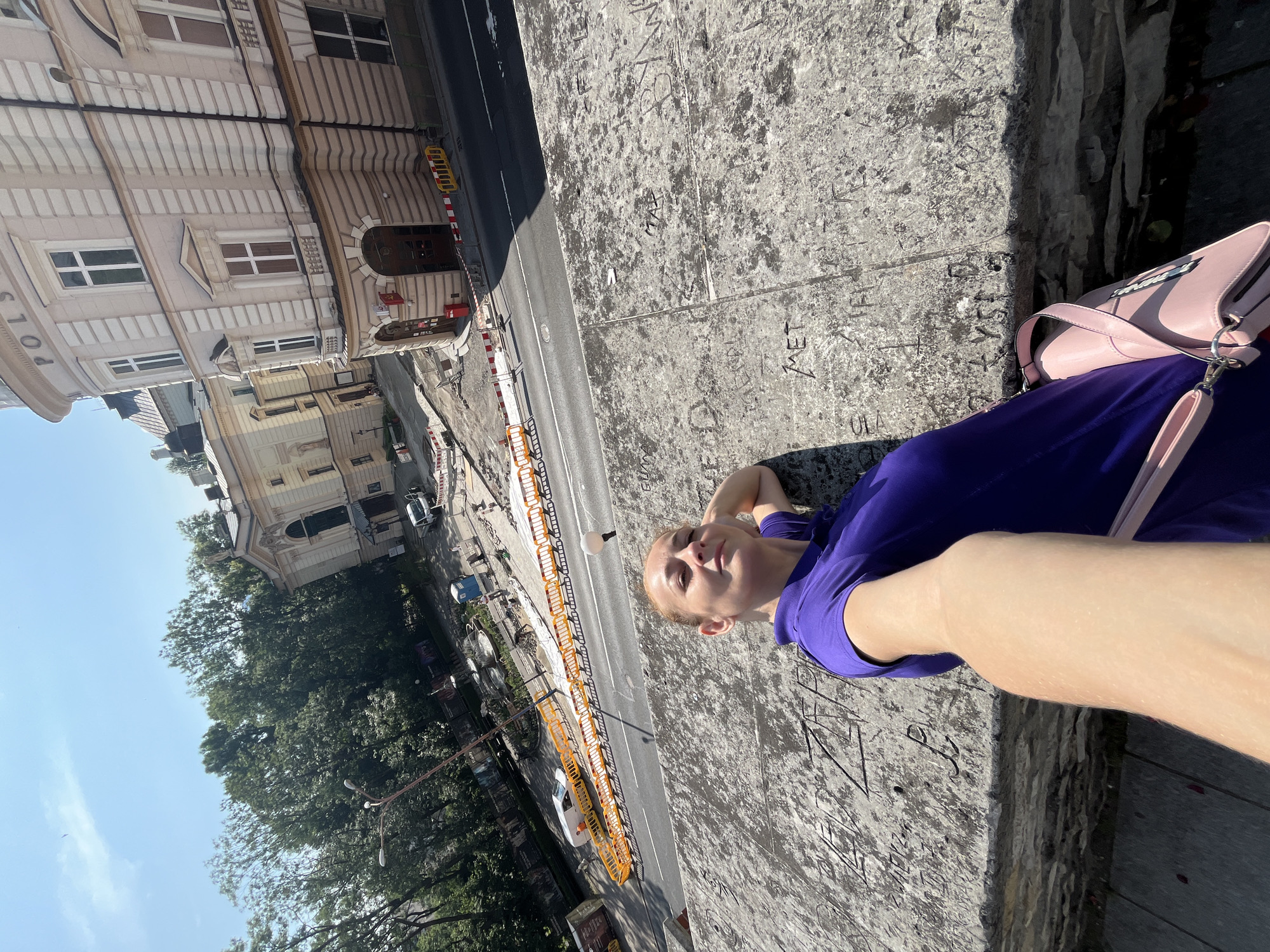 A medium shot captures a woman in a purple polo shirt taking a selfie on a sunny day. She is leaning against a concrete barrier covered in graffiti, with her right arm resting on it. A pink purse is slung over her shoulder. In the background, a street is undergoing construction with orange safety barriers and construction equipment. Beyond the construction, an ornate building with multiple windows and a curved facade stands under a clear blue sky. Trees line the street, and a lamppost is visible. The overall scene suggests an urban environment with ongoing development.