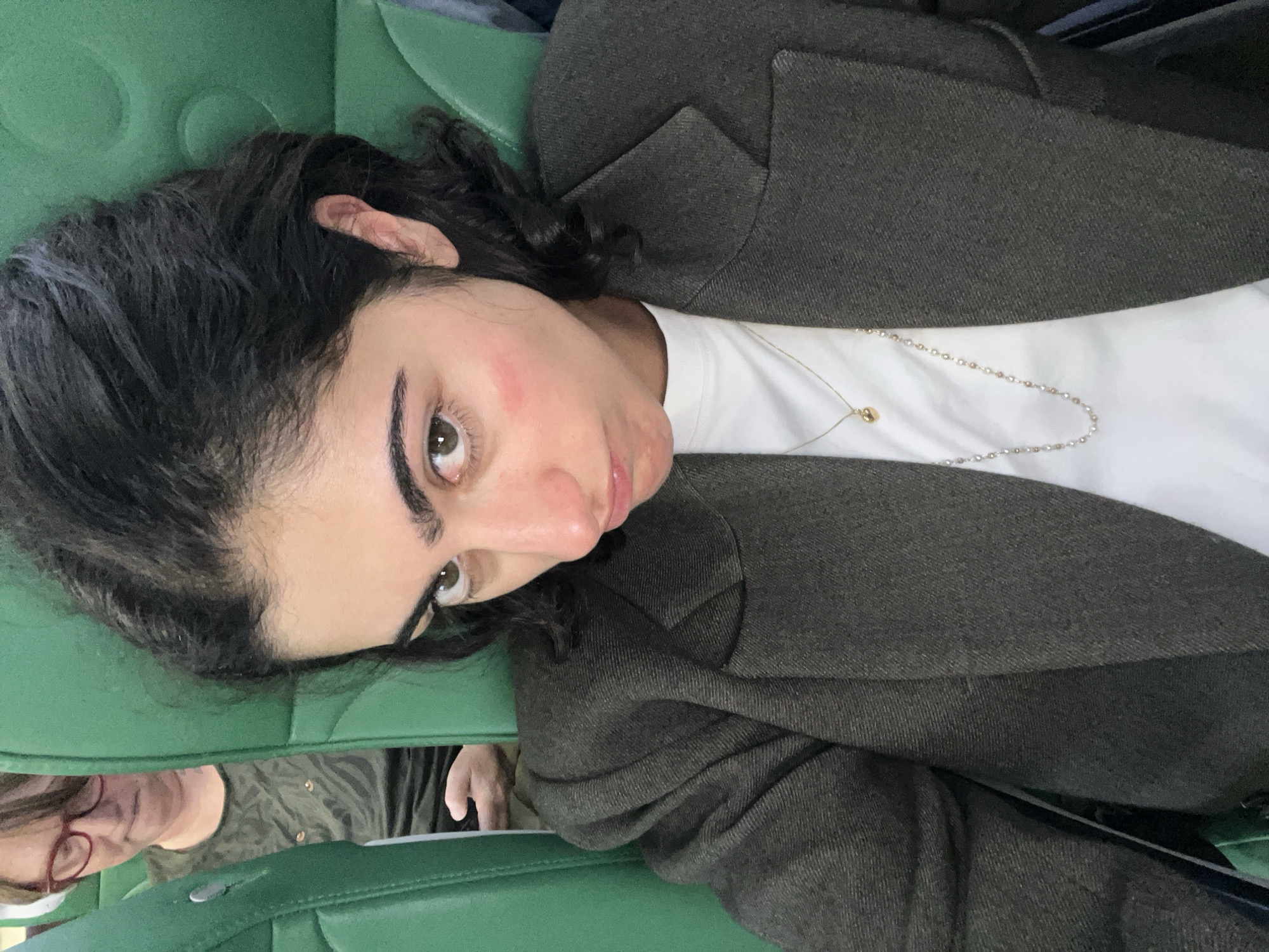 A woman with dark curly hair, wearing a dark blazer over a white t-shirt and a gold pendant necklace, lies on a green upholstered seat. The woman has light skin and prominent eyebrows. She appears to be sitting near another person with glasses, and is looking at the camera. The location is likely a public transport vehicle, potentially in or near Sant Josep de sa Talaia, Spain.