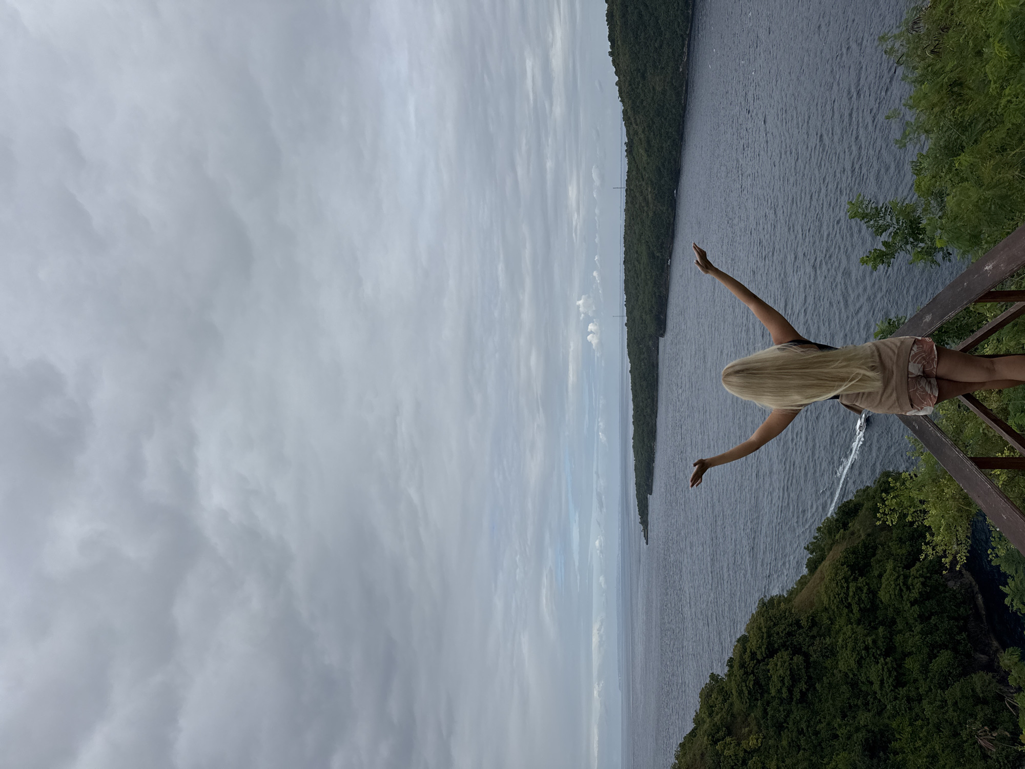 A horizontally oriented medium shot shows a woman with long blonde hair standing with her back to the viewer on a wooden platform overlooking a body of water. She has both arms raised out to the sides.
The water stretches toward a tree-covered shoreline in the distance beneath a cloudy sky. Another tree-covered landmass extends from the lower right corner.
The location is Nusa Penida, Indonesia.