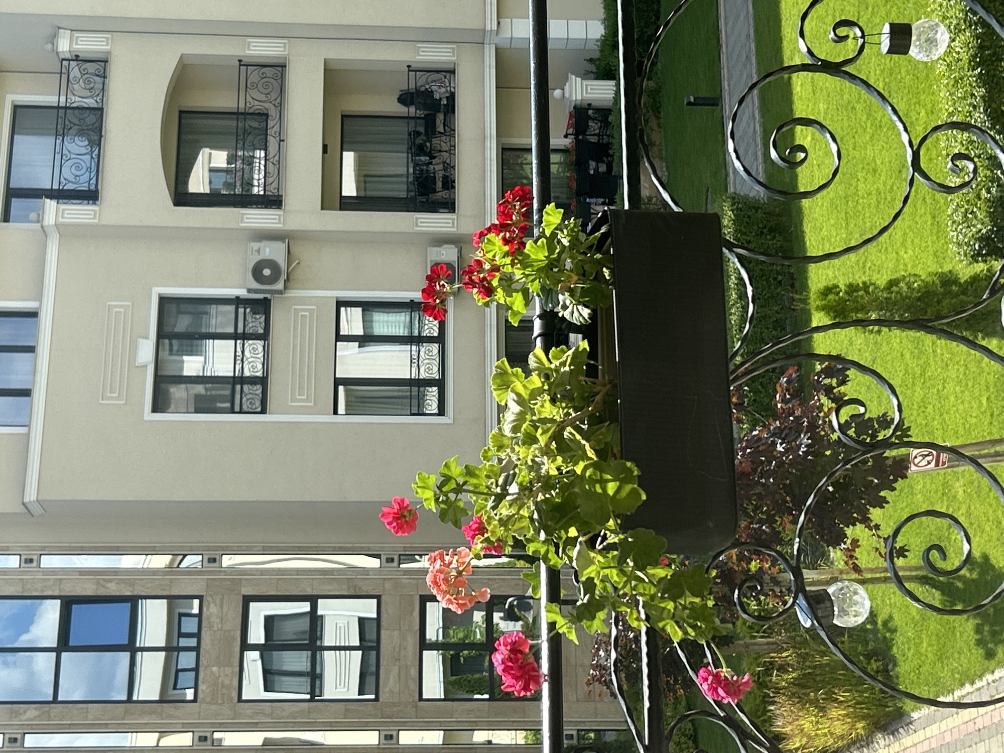 The image depicts a close-up view of a balcony railing with a flower planter in the foreground. The planter contains vibrant red and pink geraniums with lush green foliage. Behind the flowers, a black metal balcony railing with decorative scrollwork is visible, and a small solar-powered light fixture hangs from it.
In the background, a modern apartment building with beige facade and balconies with ornate railings is seen. Reflections of the sky and surrounding buildings are visible in the windows. To the left, another building with a facade of stone and glass windows is present. A green lawn, paved walkway, and some trees are visible below the balcony. The setting appears to be a sunny day.
The building's architectural style, with its ornate railings and detailed facade, is characteristic of buildings found in the Kapana Creative District of Plovdiv, Bulgaria. This district is known for its blend of historical architecture with modern renovations, housing numerous cafes, galleries, and artisan shops. The presence of the apartment building and the well-maintained greenery suggests a residential or mixed-use urban area within Plovdiv.