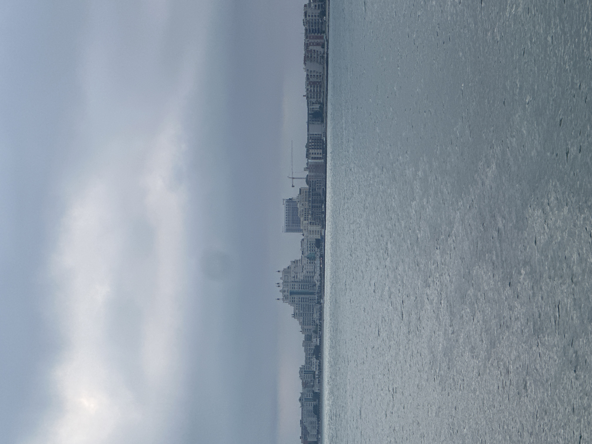 A wide shot depicts an overcast waterfront city scene with a prominent body of water in the foreground and a distant urban skyline under a grey sky. The lower two-thirds of the image are filled with a calm, rippled expanse of water, appearing greyish-blue under the muted light. Small, uneven ripples are visible across the water's surface.
The upper third of the image features a horizon line separating the water from a densely built-up urban area, topped by a heavily clouded, uniform grey sky. No direct sunlight is apparent.
The cityscape, situated along the water's edge, consists of numerous multi-story buildings. A distinctive, large, white or light-colored building with a terraced, somewhat pyramidal or conical shape and multiple domed or pointed roof structures stands out near the center-left of the skyline. To its right, other multi-story residential or commercial buildings extend along the waterfront. Two tall construction cranes are visible behind some of the buildings towards the right side of the skyline, indicating ongoing development. The buildings vary in architectural style and height, forming a continuous urban stretch. No individuals, vehicles, or specific actions are discernible within the frame. The overall atmosphere is subdued due to the overcast weather. The precise location within the city of Unknown, Unknown, cannot be determined from the image.