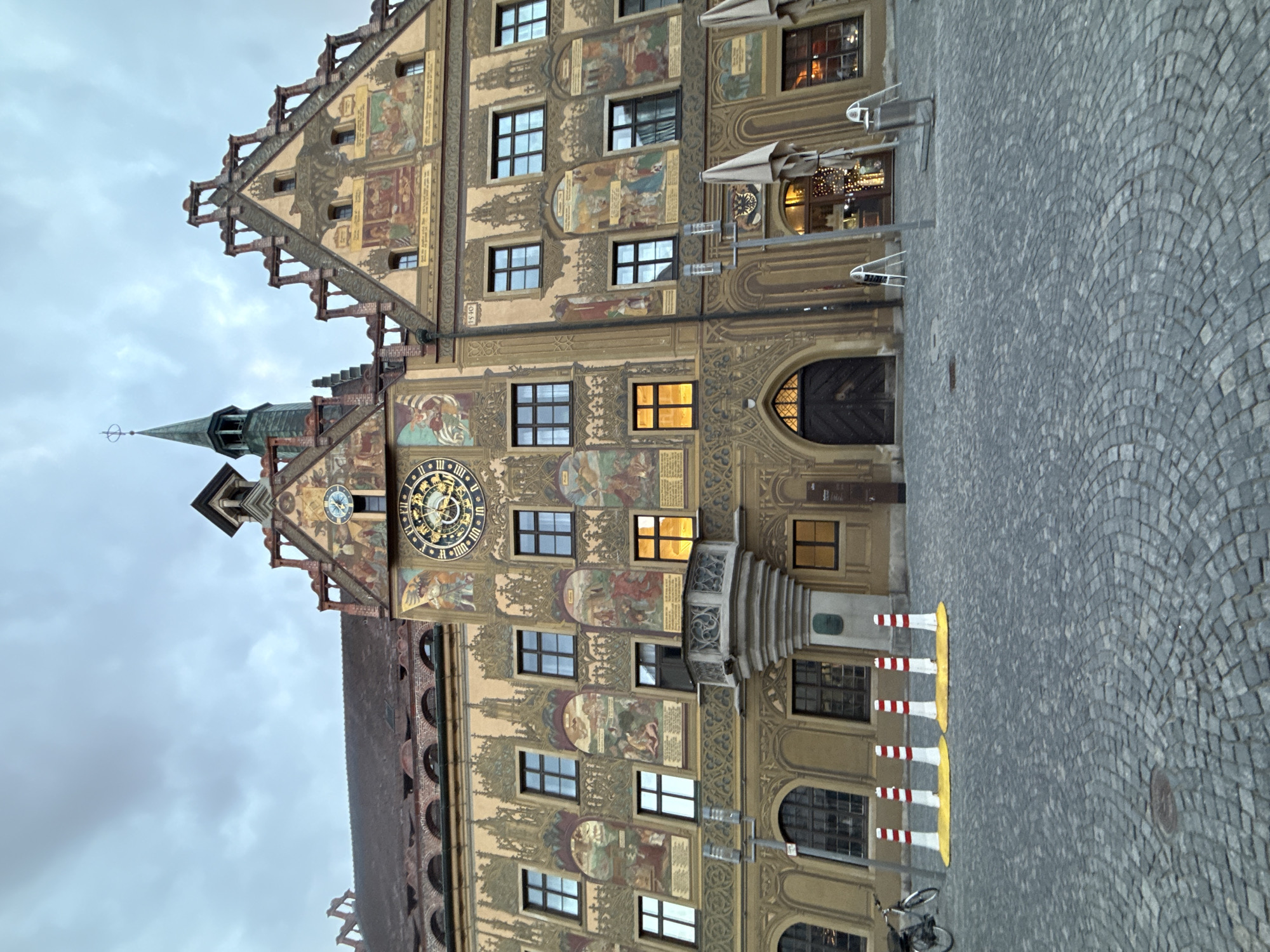 The image depicts the historical Rathaus (Town Hall) in Ulm, Germany, under an overcast sky. The building's multi-story facade is extensively decorated with numerous vibrant frescoes depicting historical and allegorical scenes, framed by ornate architectural patterns in yellow, green, and red hues. A prominent astronomical clock with gold Roman numerals and intricate mechanisms is centered on the facade, below a smaller, simpler clock face located on a higher gable to the right. A slender, green-roofed spire rises from the central roofline.
The facade features multiple rectangular windows with dark frames; some lower-level windows on the left and center-right are illuminated from within, suggesting artificial lighting. A stone oriel window with carved details projects from the facade on the left side, above a ground-level arched wooden doorway. A second arched wooden doorway is visible further to the right.
The foreground is a public square paved with grey cobblestones, arranged in a fan-like pattern. Five white and red striped bollards, each with a yellow circular base, are arranged in a linear formation on the cobblestones to the left of the center. A black bicycle is parked upright near a pole on the far left. A dark street lamp pole with multiple luminaires stands in the middle ground. To the right, two beige, folded patio umbrellas are present near a building entrance, accompanied by two white A-frame signs and a metal trash receptacle. No people are visible in the scene.