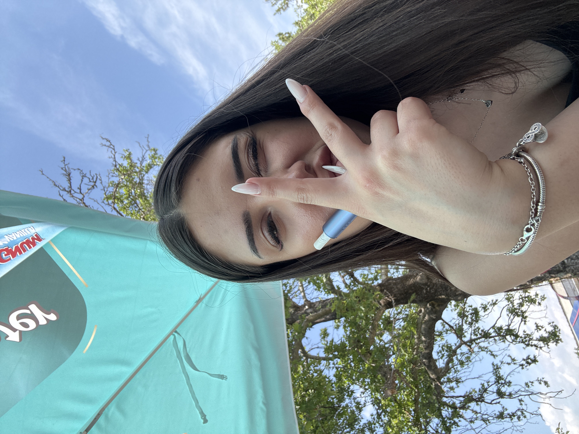 A young woman is depicted in a close-up outdoor shot, partially obscured by her left hand. She has long dark hair, visible eyelashes, and long, pointed nails with a light polish. Her left eye is closed in a wink, and she holds a blue and white electronic device between her index and middle fingers, which are configured in a "V" sign. On her left wrist, she wears a silver bracelet featuring multiple chains and charms, including an infinity symbol and a small heart. A delicate silver necklace with tiny heart pendants is visible around her neck. In the upper left background, a large turquoise umbrella or canopy displays reversed text, including "19" or "91" and a portion of "TEMA," along with gold stripes. Behind the woman and the umbrella, a large deciduous tree with green foliage and prominent branches stands against a bright blue sky with scattered white clouds, indicating an outdoor setting in Kostievo, Bulgaria.