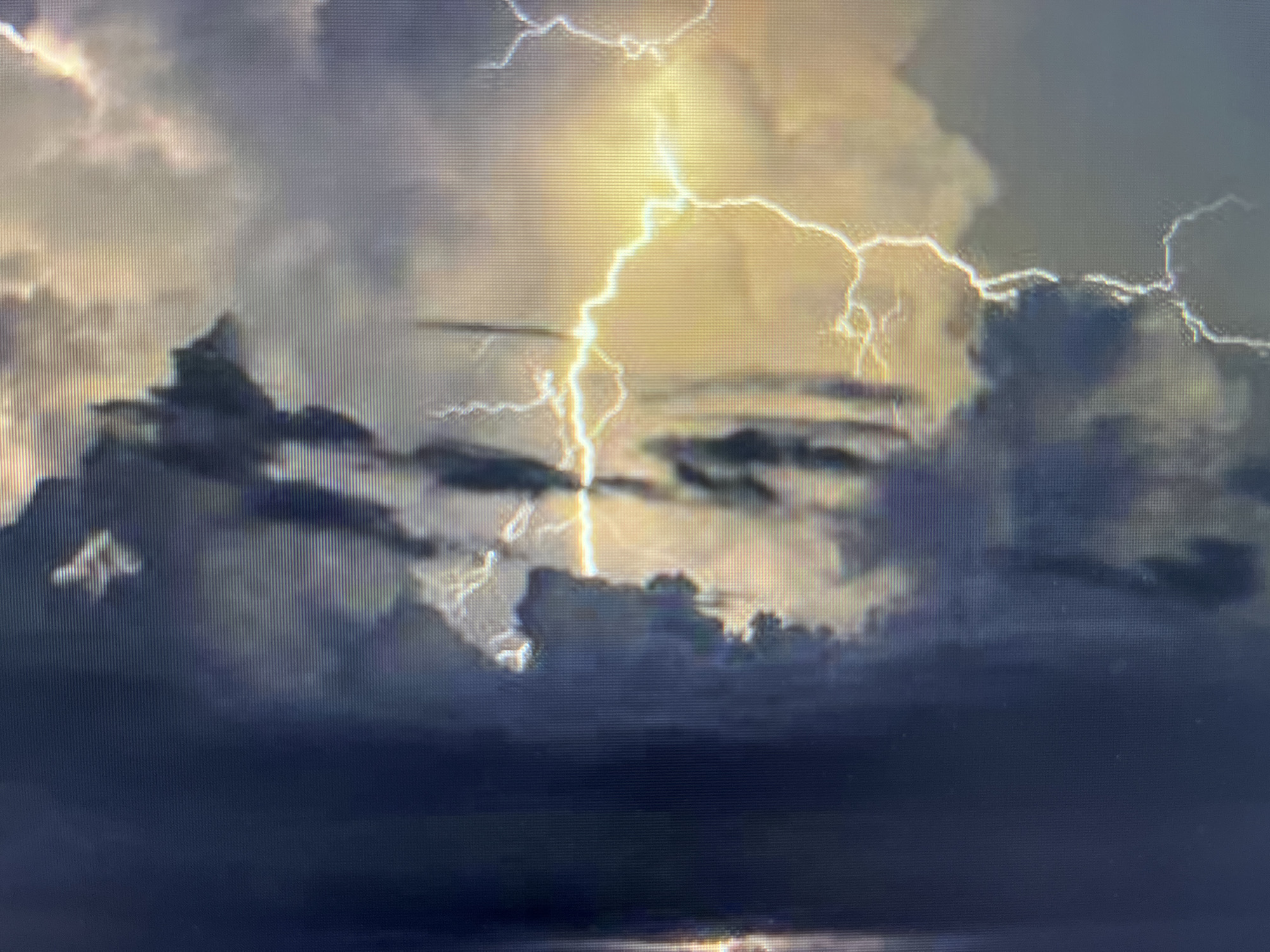 The image depicts a dramatic nighttime or twilight thunderstorm, showcasing a turbulent sky filled with dark, heavy clouds illuminated by powerful lightning strikes. A prominent, bright yellow-white lightning bolt descends vertically in the upper center, branching out with numerous smaller, intense flashes across the sky. The light from the lightning illuminates the edges and interiors of the dense cloud formations, creating a striking contrast between the bright flashes and the deep, somber tones of the storm clouds.
In the lower section of the image, a dark, reflective surface, likely a body of water or wet ground, can be seen, with subtle reflections of the distant lightning. On the horizon or within the cloud structure, particularly visible in one of the close-up crops, there appears to be a silhouette resembling a mountain peak or a distinctive cloud formation.
The activity captured is an intense lightning storm. The weather is severe, characterized by the dark clouds and frequent electrical discharges. There are no people or explicit man-made objects visible. The overall mood is powerful and awe-inspiring, highlighting the raw energy of nature. The image displays a distinct pixel grid pattern, suggesting it is a photograph of a digital screen. Given the location context of Varna, Bulgaria, the scene likely portrays a storm over the Black Sea or the coastal landscape. No visible text is present in the image.