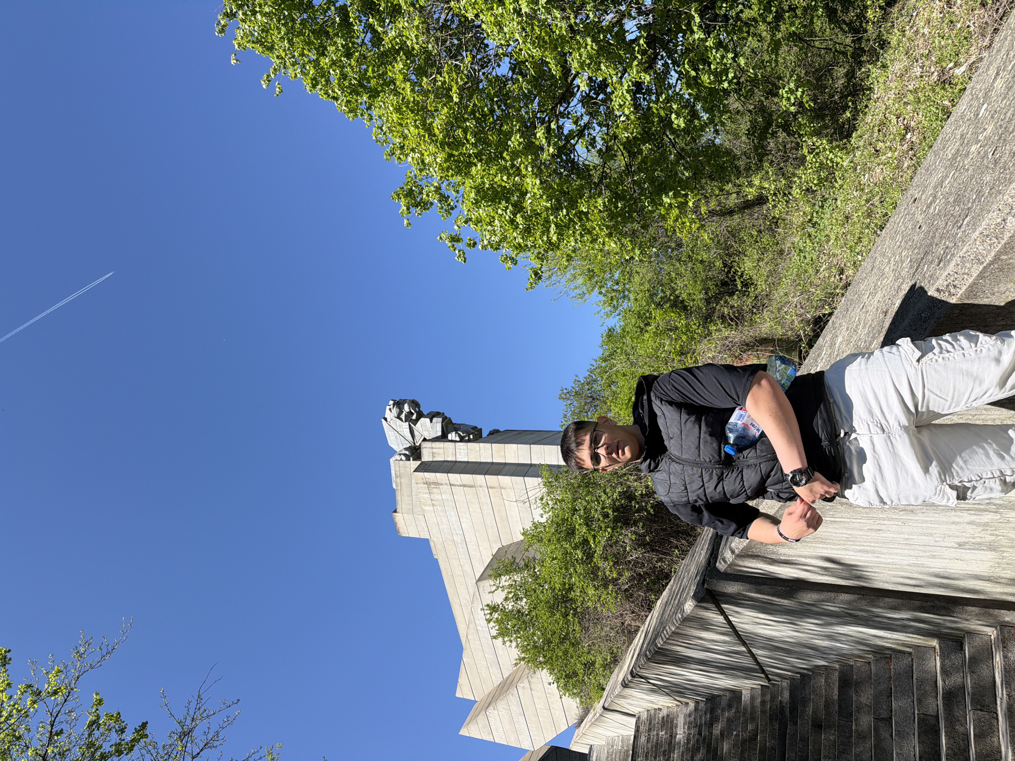A man wearing a black jacket and white pants is photographed from a slightly low angle, reclining on a concrete structure. In the background is the Founders of the Bulgarian State Monument in Shumen, Bulgaria. The sky is blue and a contrail can be seen in the upper left portion of the image. Trees with green leaves are visible behind and beside the monument. The man is wearing a watch on his left wrist and a bracelet on his right. He appears to be looking directly at the camera. A water bottle is next to him. The concrete structure on which he is lying appears to have a staircase.