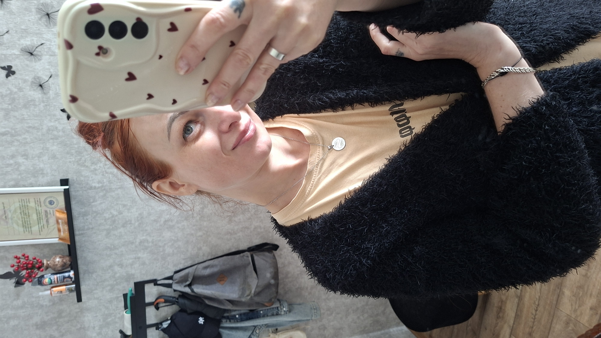A woman with red hair pulled back in a bun, wearing a tan t-shirt and a fuzzy black cardigan, takes a mirror selfie. She is wearing a silver necklace with a circular pendant and a silver ring on her right index finger. A silver bracelet adorns her left wrist. Her phone case has a pattern of small red hearts. In the background, a shelf holds a vase with red berries, a framed document, and a small chocolate bar. A rack with several bags and jackets is also visible. The wall has a textured gray finish with some decorative butterfly and dandelion designs. The floor is made of wooden planks. The scene appears to be indoors, possibly in an apartment, in the city of Myadzyel, Belarus.