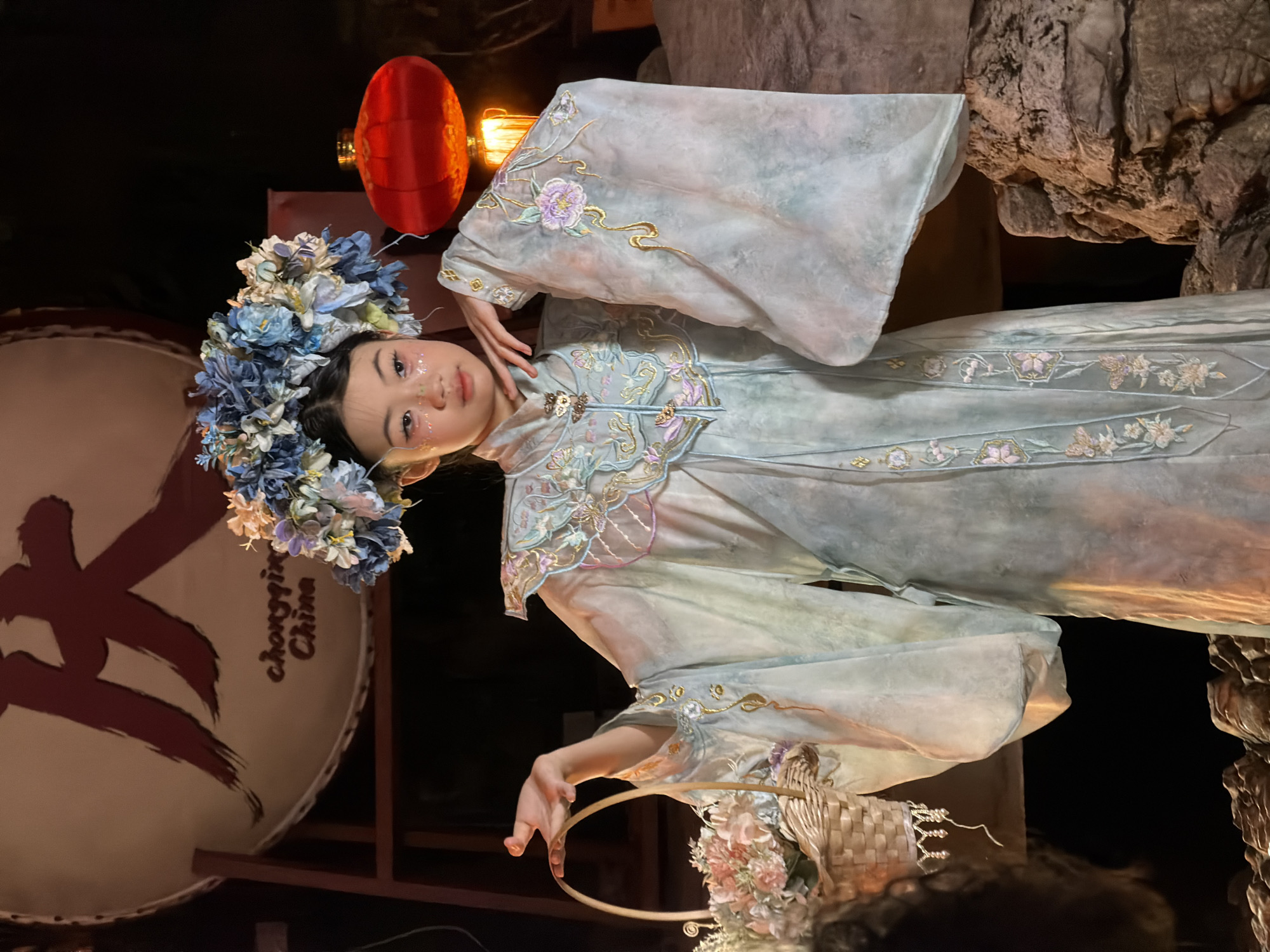 A young East Asian girl is dressed in a traditional pale blue and green Chinese garment with intricate floral embroidery. She wears a large, elaborate floral crown in shades of blue and white. Her face is adorned with subtle glitter makeup. She holds a woven basket filled with flowers in her left hand, positioned slightly in front of her. Her right hand is raised near her face. In the background, a large, circular object with Chinese calligraphy is visible, along with a red lantern and decorative rocks. The scene appears to be a staged portrait. The backdrop suggests a setting in Yuzhong District, China, potentially related to a cultural or promotional event, given the calligraphy and lantern. The lighting is warm and dramatic, highlighting the subject.