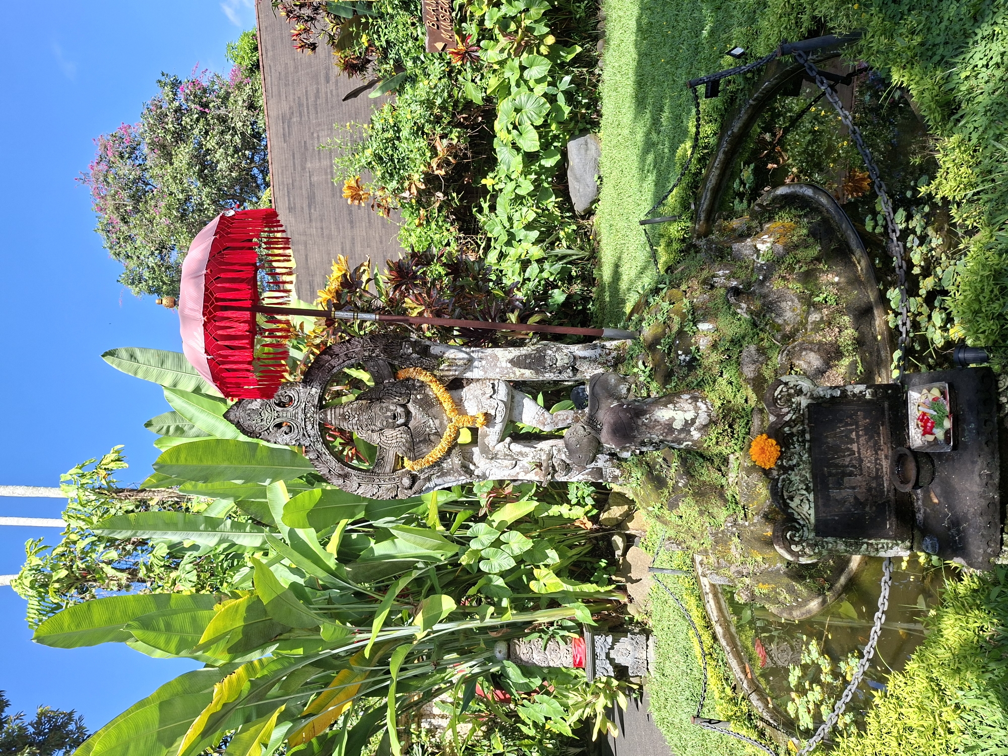 The image shows an outdoor scene in the Ubud District, Indonesia, featuring a Balinese statue adorned with a yellow garland and a red umbrella. Lush tropical plants surround the statue, including large banana leaves. Behind the statue, there's a structure with a dark wooden facade, and a tree with pink flowers is visible in the background under a bright blue sky. To the statue's right, there is a stone structure with a small pond at the base and a metal railing around it. On top of the structure are offerings, including an orange flower and a dish with colorful items.