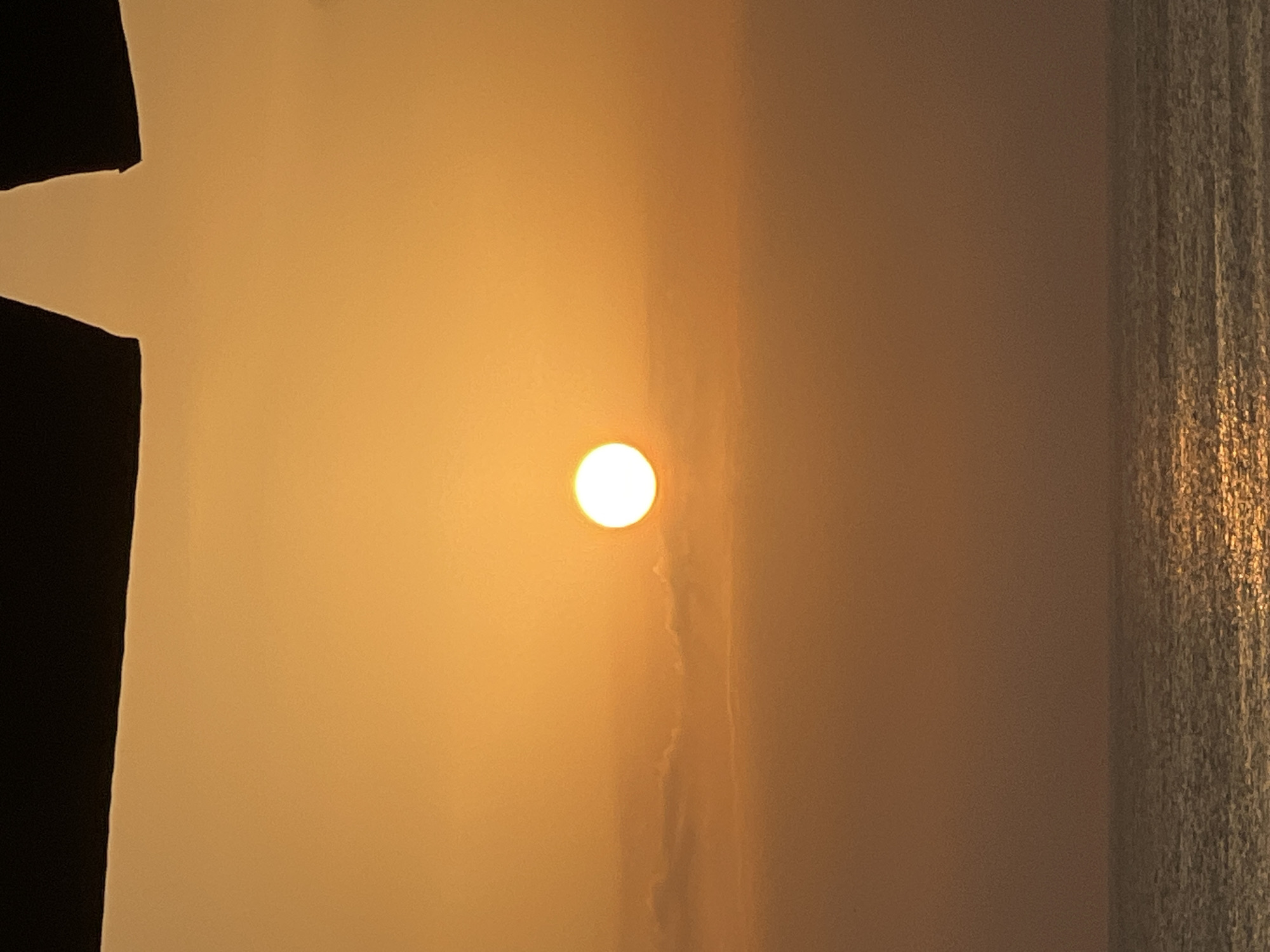 The image shows a sunset over the ocean in Kuta, Indonesia. The sun is a bright orange orb, partially obscured by faint clouds. The sky is a gradient of orange and yellow, with darker, amorphous shapes silhouetted at the top. Below the sky, the ocean stretches to the horizon, with its surface reflecting the warm hues of the sunset. The water appears calm, with gentle ripples catching the light. There are no people or man-made structures visible in the scene.