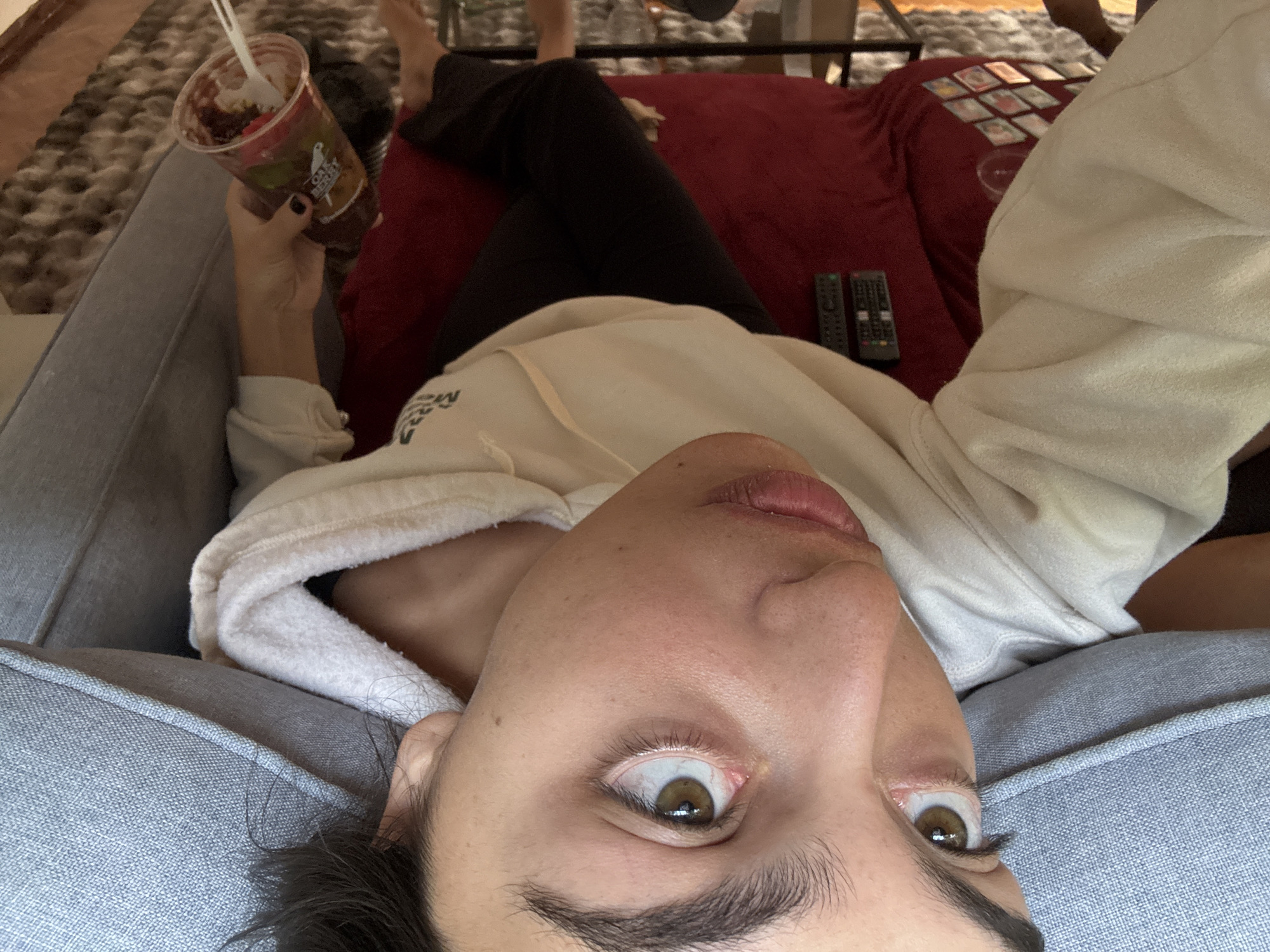 The image is a first-person perspective, likely a selfie, of a person relaxing indoors. The subject lies on a grey upholstered couch or chair, looking directly up at the camera. Their face, featuring wide brown eyes and slightly parted lips, dominates the foreground. They are wearing a light-colored, possibly off-white, hooded sweatshirt and dark pants. Their left hand holds a plastic cup with a lid and a white spoon, filled with a dark food item; the cup bears an "OAKBERRY" logo. Dark nail polish is visible on their fingers.
In the background, the subject's legs are crossed and rest on a red blanket or throw that covers another piece of furniture. On this red surface, two remote controls are placed to the left, and several small, rectangular cards, possibly tarot cards or photos, are fanned out next to a clear round object. A patterned rug is visible on the floor, and parts of another person's feet and a metal furniture frame are in the out-of-focus background.
The scene depicts a casual, relaxed atmosphere, likely in a living room or bedroom in Mexico City during the daytime. The person appears to be enjoying a snack while possibly watching TV or engaging in card-related activities. The wide-eyed gaze of the subject creates a direct connection with the viewer.
