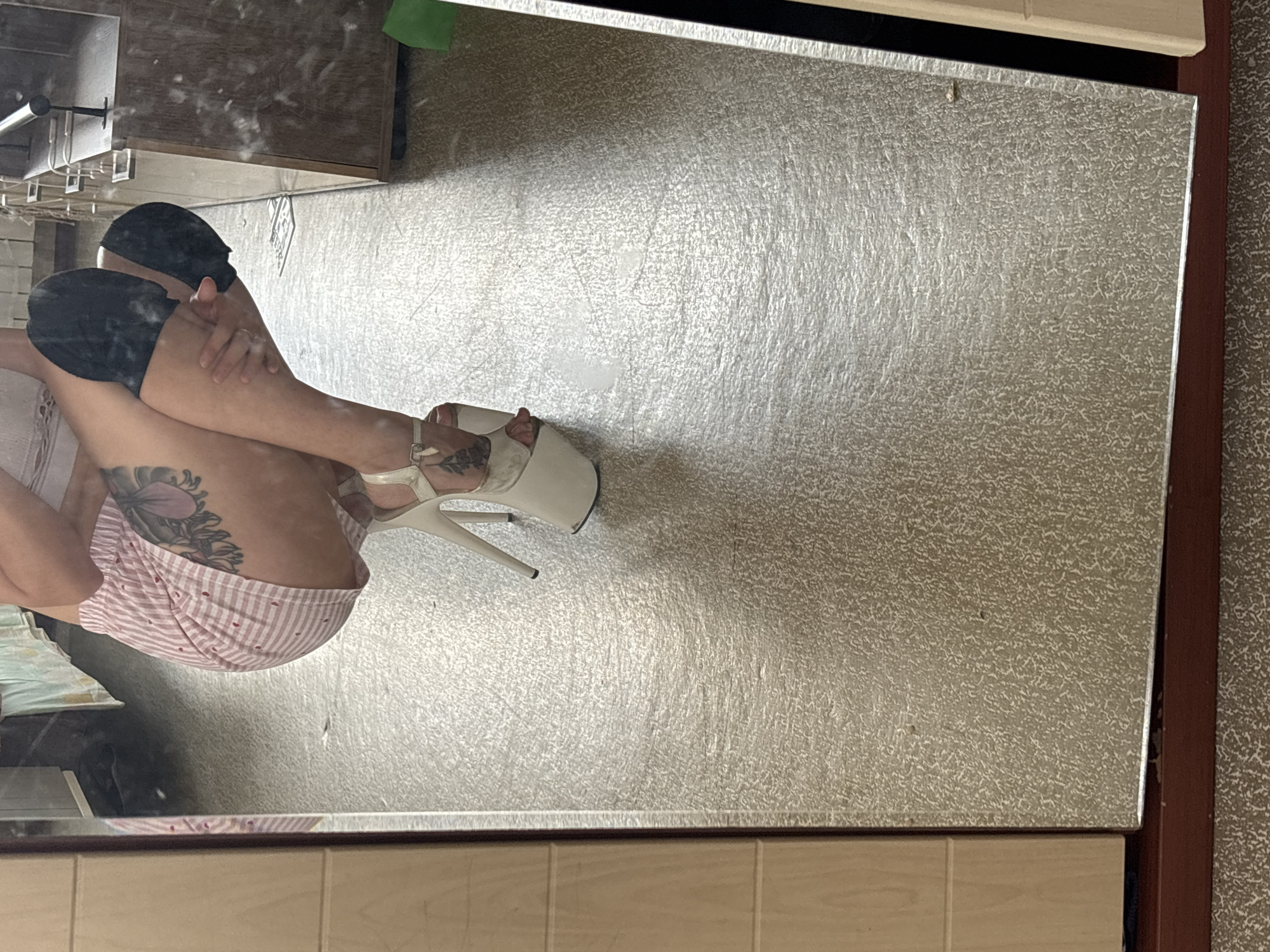 A person is crouching down, reflected in a full-length mirror. They are wearing a pink and white striped skirt, a white top with lace detail, and white platform heels. The person has tattoos on their leg. The background is a room with a textured floor and what appears to be shelves or furniture. The mirror reflects a portion of the room, including part of a shelf unit and a green object on the floor. The image does not provide any information about the location within the city of Naberezhnye Chelny, Russia.