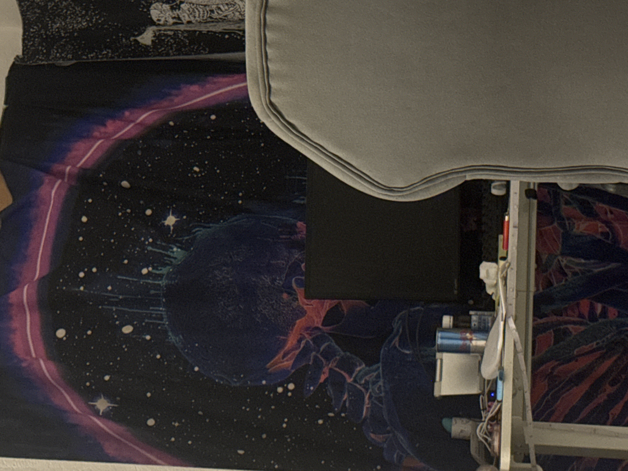 A gray office chair with a textured headrest is positioned in front of a desk cluttered with various items. Behind the desk, a tapestry with a cosmic or psychedelic design featuring a skeletal figure and stars covers the wall. A black computer monitor is visible on the desk, partially obscured by the chair. The overall scene suggests a personal workspace, possibly in a bedroom or office. The location is unknown.