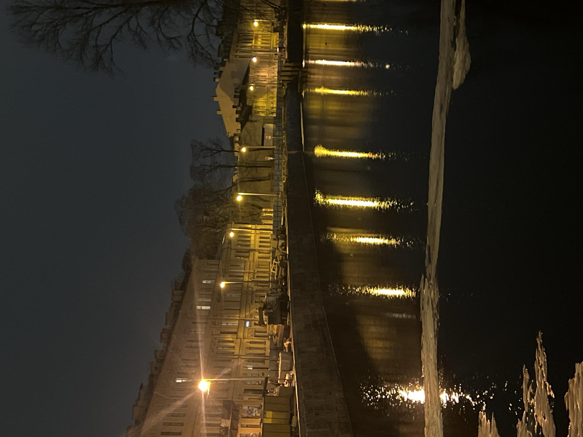A nighttime image displays a canal in the historic center of Saint Petersburg, Russia, likely during winter or early spring. The environment is illuminated by numerous streetlights emitting warm, yellow-orange light, which creates prominent, elongated reflections on the dark canal water. Irregular patches of white ice or slush are visible floating on the water's surface, particularly in the foreground.
The far bank features a stone-clad embankment. Along the top of this embankment, a series of tall, pole-mounted streetlights are spaced. The embankment appears to be a work or storage area, containing several large, yellow rectangular containers or barriers, stacked concrete blocks, and what appears to be a large utility vehicle or truck situated on the left-center of the embankment.
Behind the embankment, a continuous row of multi-story buildings, characterized by their classical architectural style with numerous evenly spaced windows, extends across the frame. These buildings are partially illuminated by the streetlights and internal lights visible in some windows, appearing in shades of yellow and beige. Several bare deciduous trees, devoid of leaves, are visible against the dark, overcast sky behind the buildings, particularly on the far right and in gaps between structures. No individuals are visible within the scene.