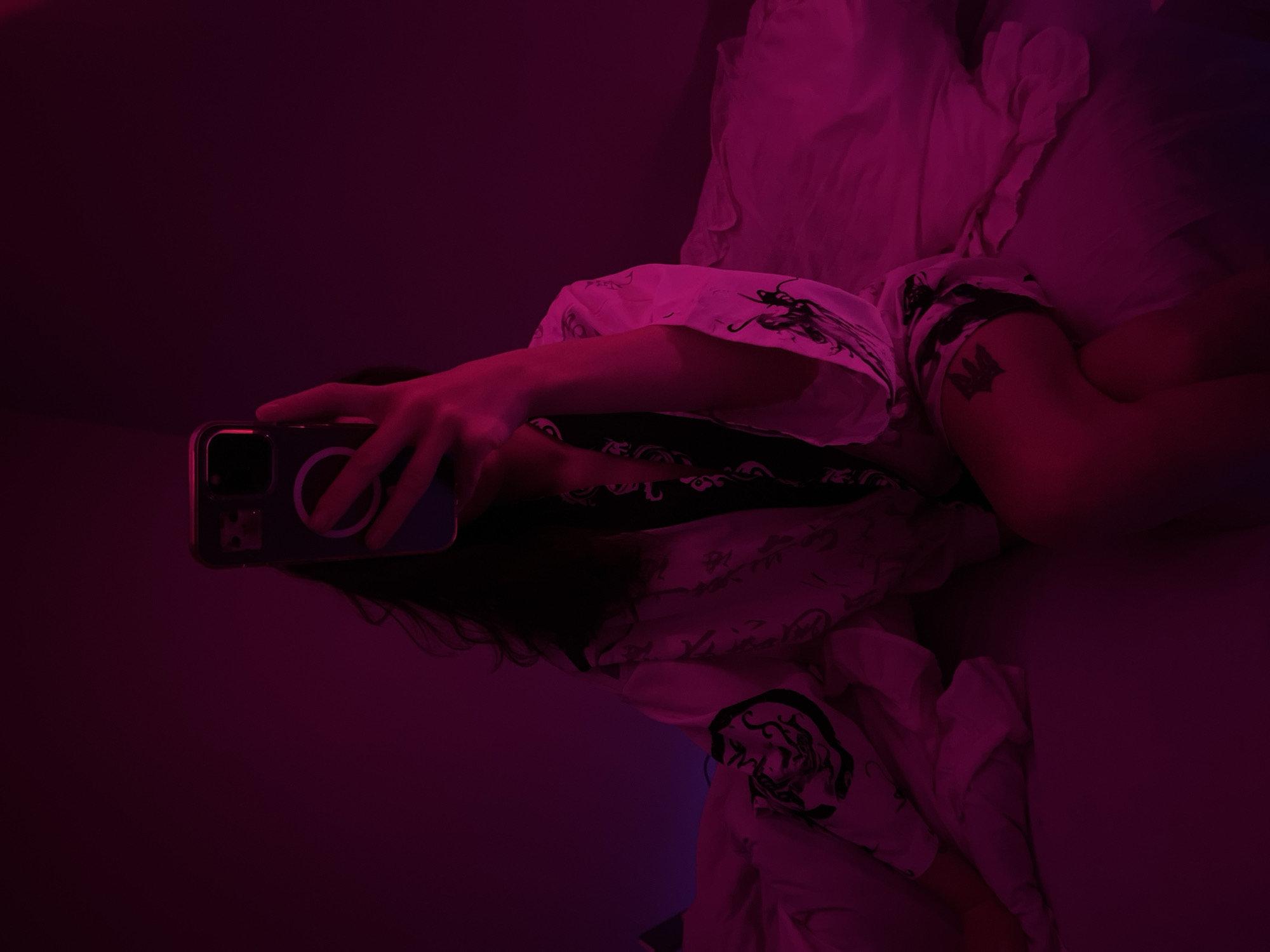 The image shows a person sitting on a bed, holding a phone to take a selfie. The person is wearing a kimono with black and white patterns. The room is dimly lit with pink and purple lighting. There is a tattoo on the person's thigh. The location of the image is not specified and cannot be determined from the image itself, and therefore, it cannot be said to be in Ternopil, Ukraine.