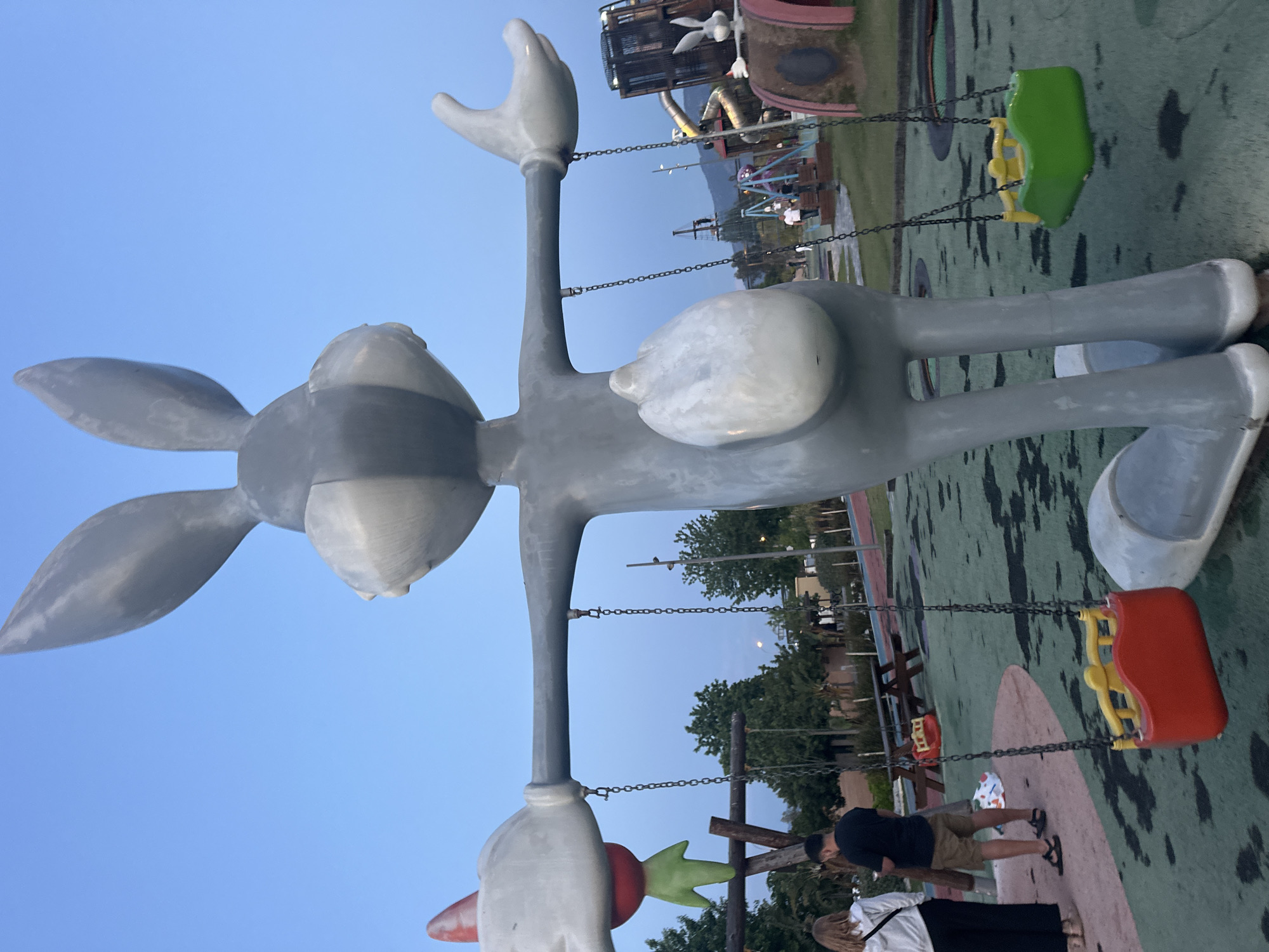 The image depicts a large, gray statue of Bugs Bunny, positioned in a park or playground. The statue has its arms outstretched and appears to be holding or supporting swings.
In the background, other play structures are visible, as well as people. The sky is blue and suggests the photo was taken in the late afternoon. The location is Fethiye, Turkey.