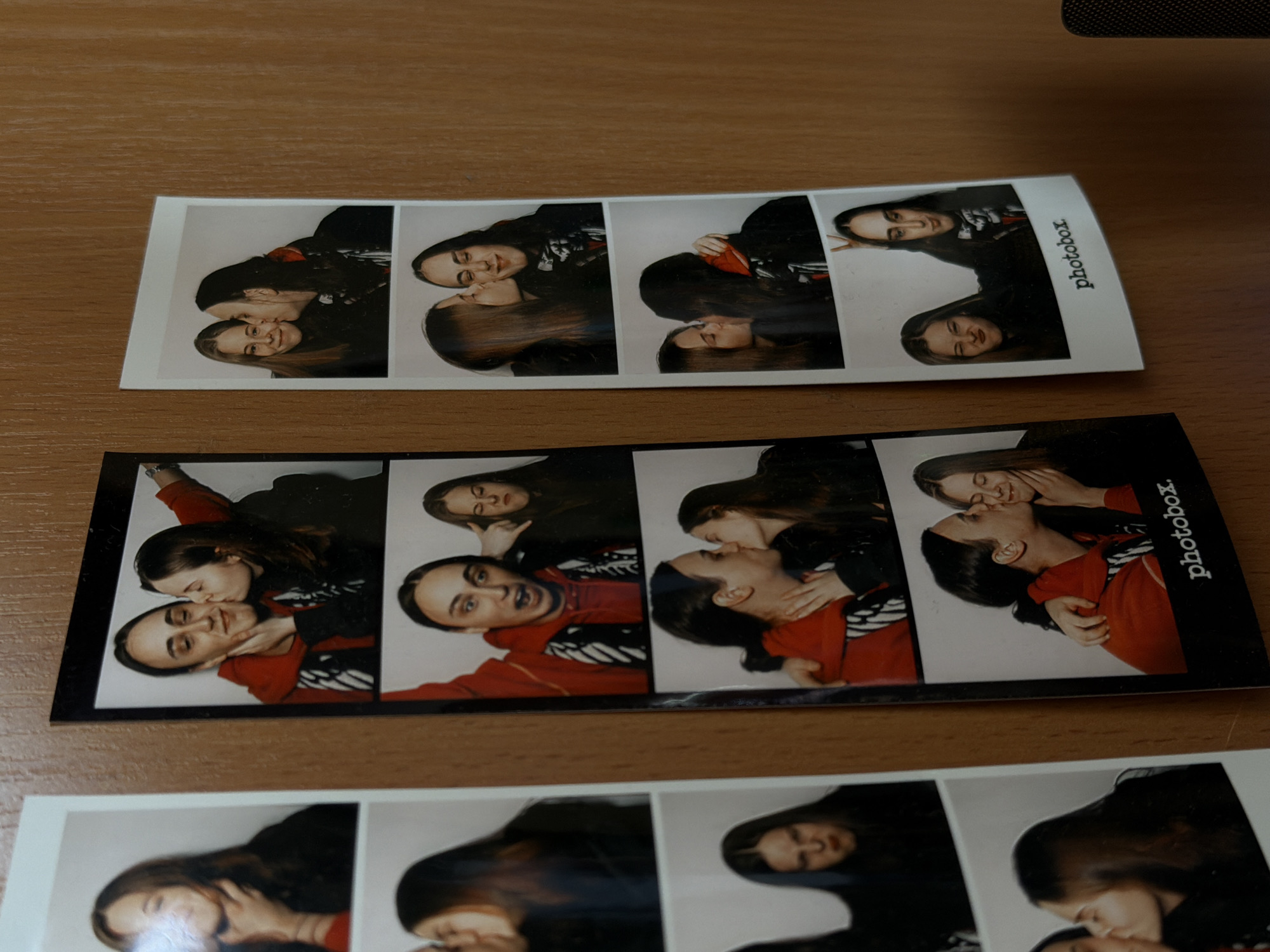 On a wooden surface, three photobooth strips are displayed. Two strips are fully visible, and a portion of the third strip is seen at the bottom of the frame. The strips are rectangular and feature multiple sequential images of two individuals, presumably a couple. The individuals are depicted in close proximity, engaged in affectionate poses such as kissing and embracing. The strips have a black border and include the word "photobox" printed on the side. The images are predominantly in black and white, though some strips show color variations due to lighting or film processing. The surface appears to be a wooden table, with a neutral tone and visible wood grain.