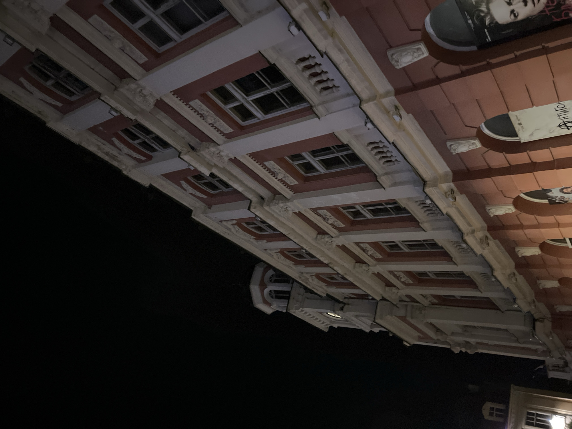 This is a low-angle, nighttime photograph of a grand, old building with a pink facade and white decorative elements. The building has multiple stories, with rows of windows visible on each level. The windows appear to have multiple panes. There are decorative moldings and columns on the facade. A tower with arched windows is visible on the left side of the building. The sky is dark and appears to be cloudless. To the lower right of the main building, there are two arched entrances on the ground floor, each with posters displayed. One poster features a close-up of a woman's face. Another poster has text that appears to be in a non-English script and includes a symbol that resembles a circled 'A'. The building is illuminated by artificial light, casting shadows. A smaller, lighter-colored building with a single window is visible to the far left of the frame.