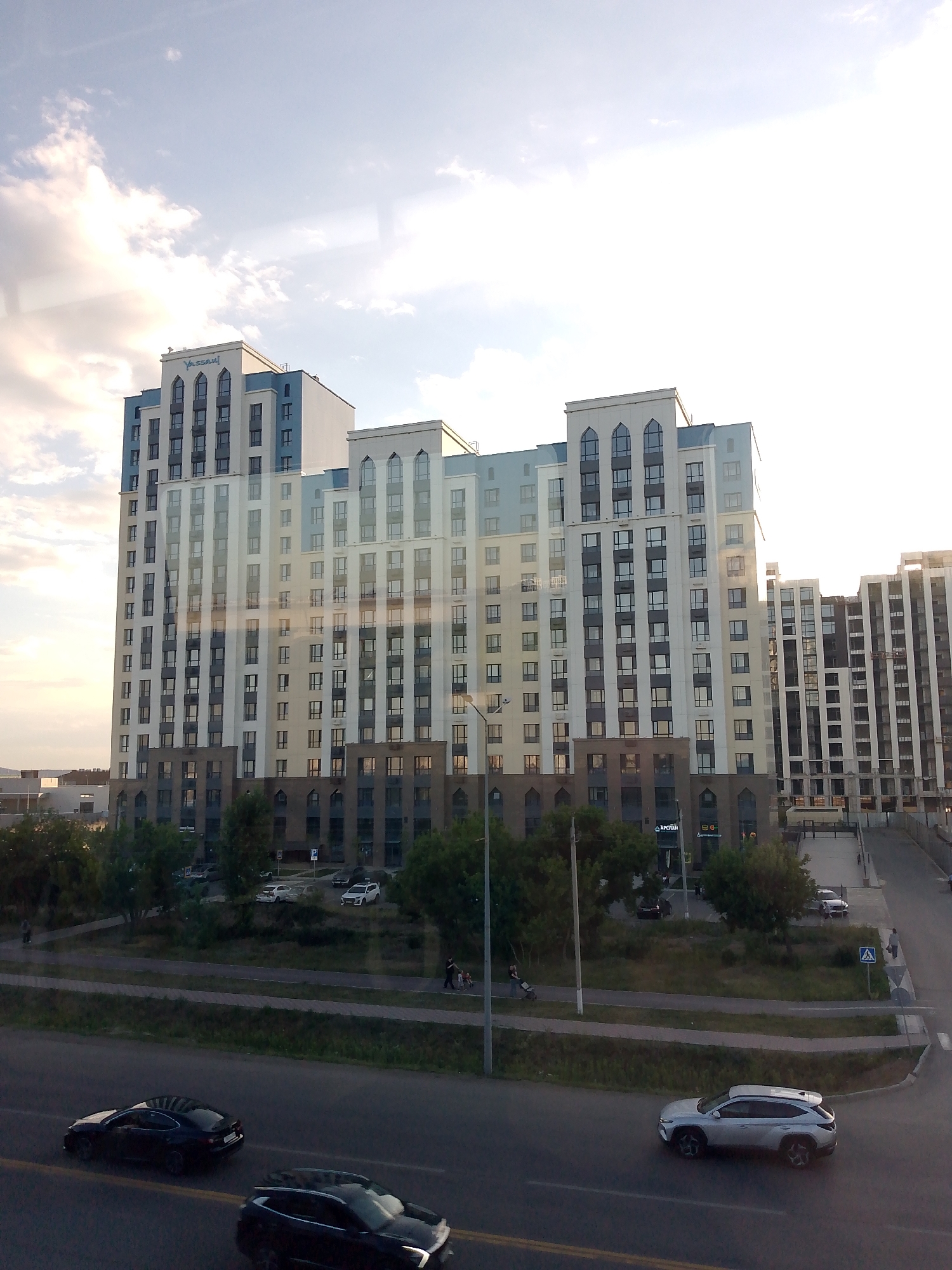 The image captures a view of a modern, multi-story apartment building with a light blue and white facade, featuring numerous windows and a distinctive arched design at the top. The building is situated in an urban environment, with other buildings and construction visible in the background. In the foreground, a road with traffic is visible, with several cars driving on it. The scene appears to be taking place during the daytime, with a bright sky and clouds indicating fair weather. The lighting suggests it might be late afternoon or early morning. There are trees and greenery interspersed between the buildings and along the roadside, suggesting a planned urban landscape. Some people are visible walking on a sidewalk or pathway next to the road.
The primary subjects are the large residential building and the road with vehicles. Objects include cars, a lamppost, and traffic signs. The scene is an urban street with residential and possibly commercial buildings.
The activity is general urban life, with cars moving on the road and people engaged in pedestrian activities. There are no specific events immediately apparent.
Notable details include the architecture of the apartment building, the presence of construction in the background, and the mix of greenery and infrastructure. The sky is bright with scattered clouds, and the sun's glare suggests it is a sunny day. There is visible text on the apartment building, which reads "YASSAU" in blue letters. Additionally, there is text on a sign on one of the buildings in the middle ground, which appears to be in Cyrillic script, possibly reading "АПТЕКА" (Apteka, meaning Pharmacy).