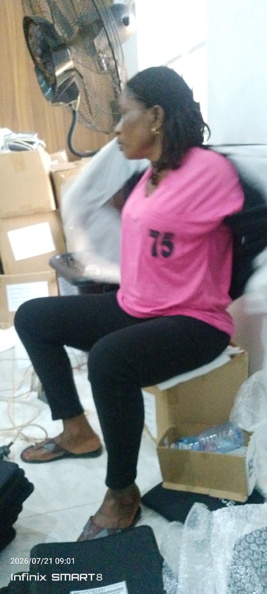In a room with beige wooden paneling, a woman is seated on a cardboard box. She is wearing a bright pink t-shirt with the number "75" printed in black on the front, and black leggings. She has dark hair and is wearing gold hoop earrings. Her feet are clad in brown and silver sandals. In the background, a black oscillating fan is mounted on the wall. Several cardboard boxes are stacked around the room, and a clear plastic bottle of water is visible inside one of the boxes. The floor is tiled and there are electrical wires visible. A timestamp in the lower left corner reads "2026/07/21 09:01" and "Infinix SMART 8". The scene appears to be indoors, possibly a storage area or office space, in Kakau, Nigeria.